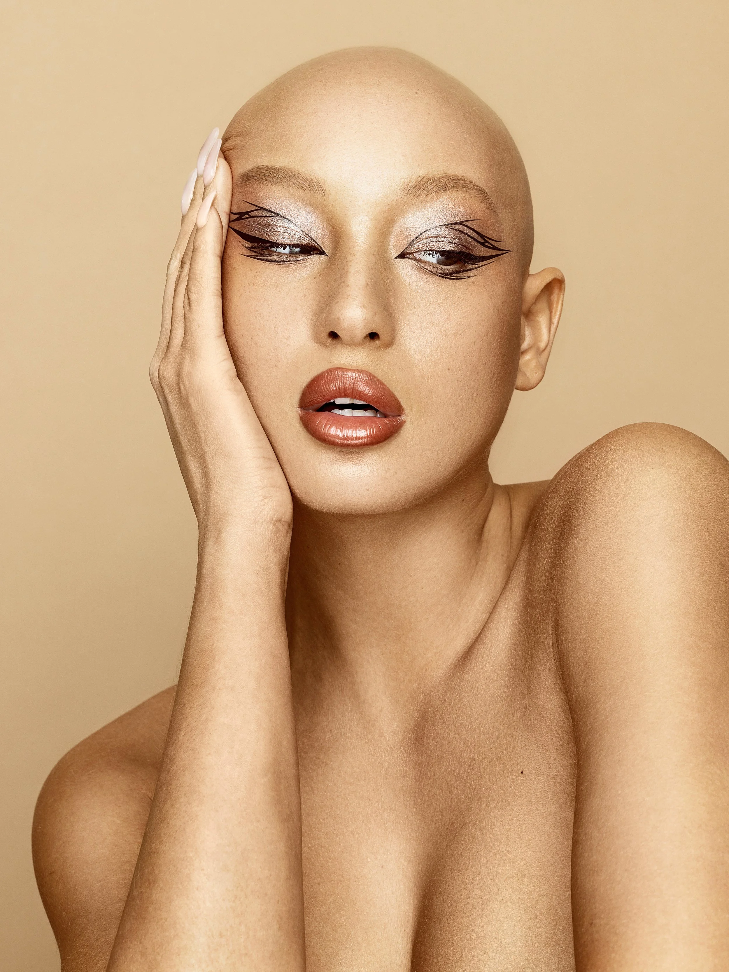 Close-up of a woman with bald head, bold makeup, and hand resting on her face, against a beige background. Beauty Photographer Los Angeles.