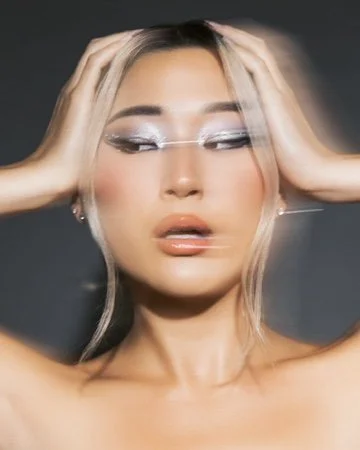 A woman with platinum blonde hair and makeup, holding her head with both hands, looking sideways, against a dark background. Beauty Photographer Los Angeles.