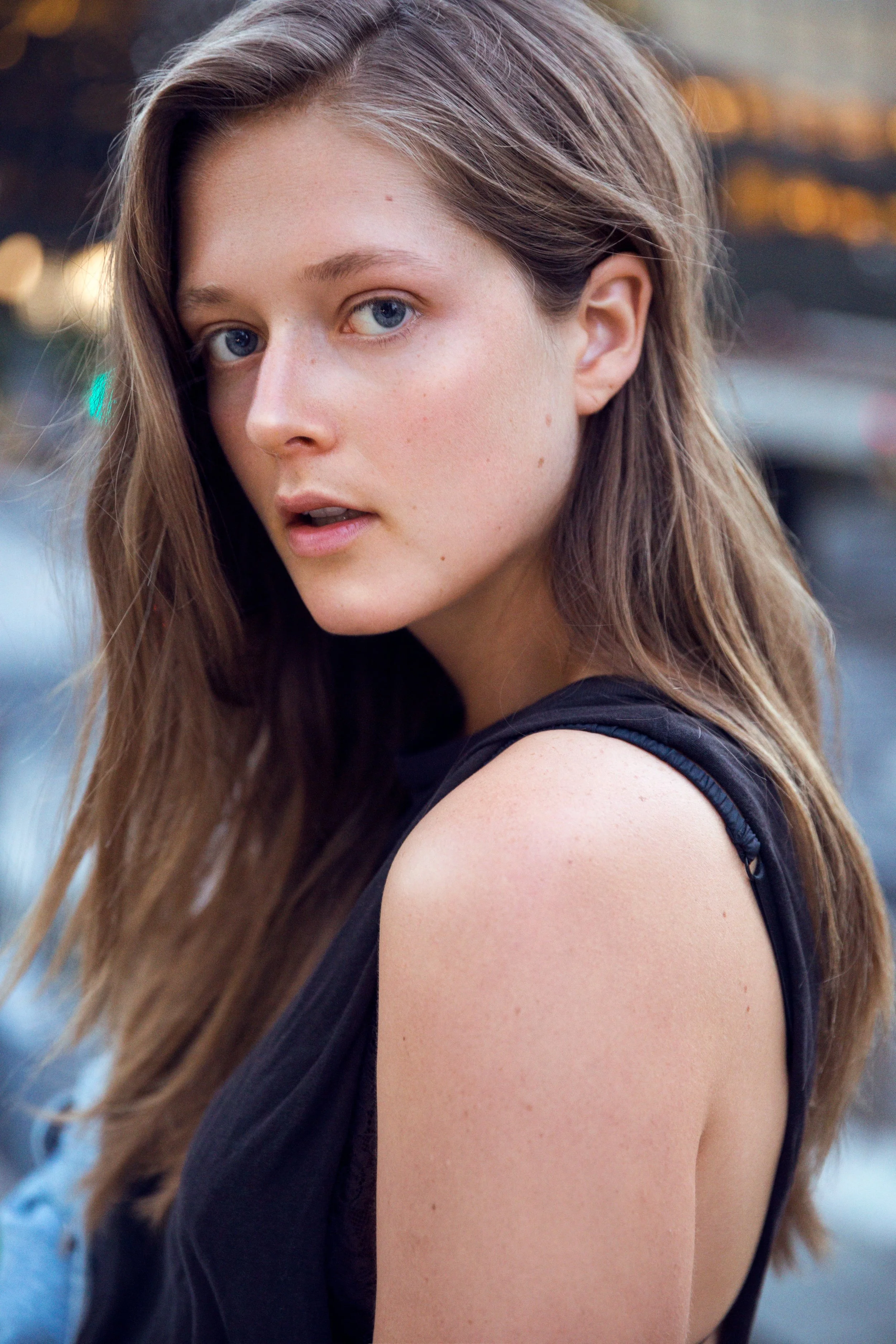 Close-up of a young woman with long brown hair and blue eyes, wearing a black sleeveless top, outdoors during daytime.