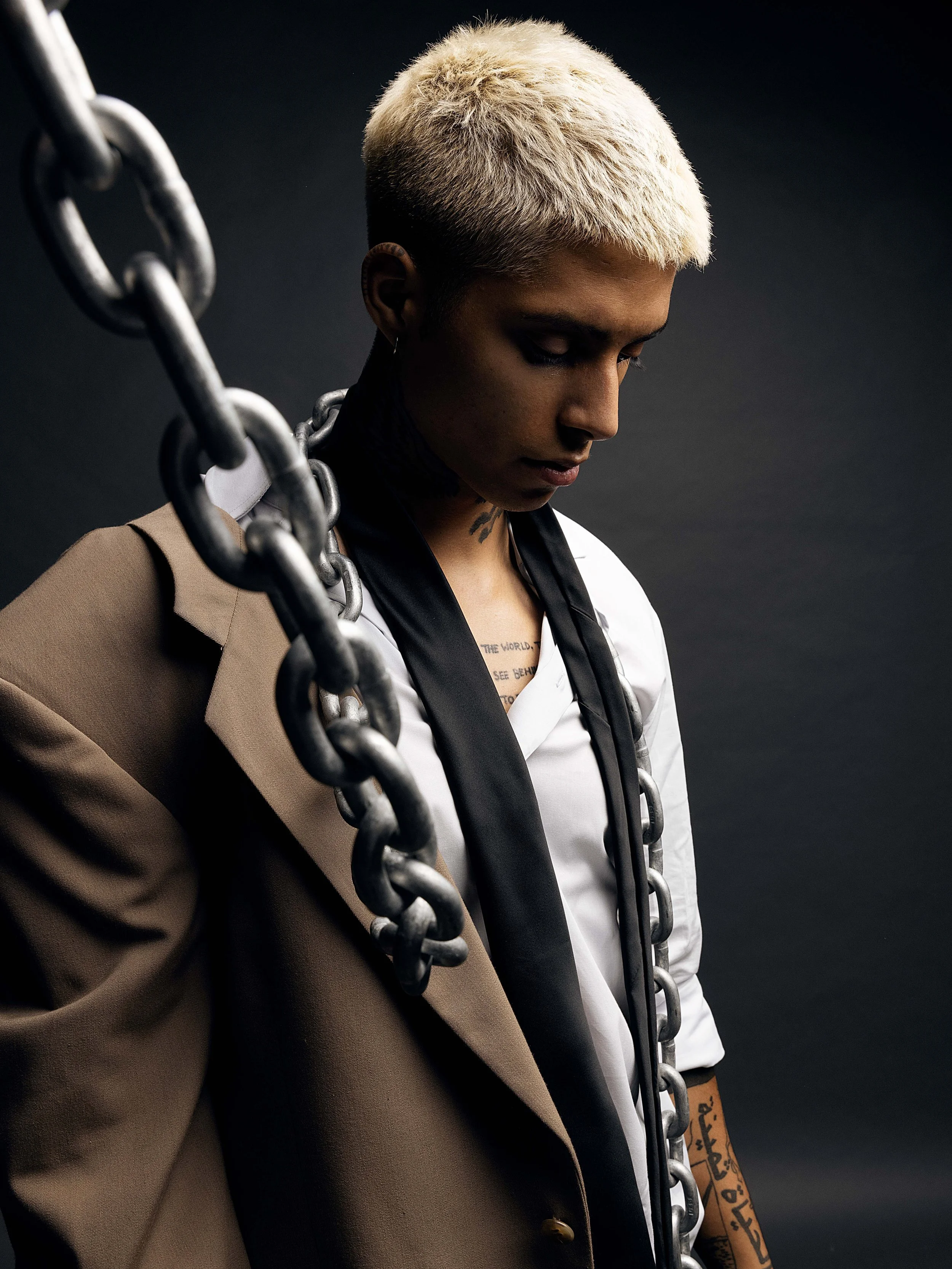 Person with short, bleached blonde hair, wearing a black tie, white shirt, tan blazer, and jewelry, with a large chain of metal links draped over their shoulder and neck, against a dark background.