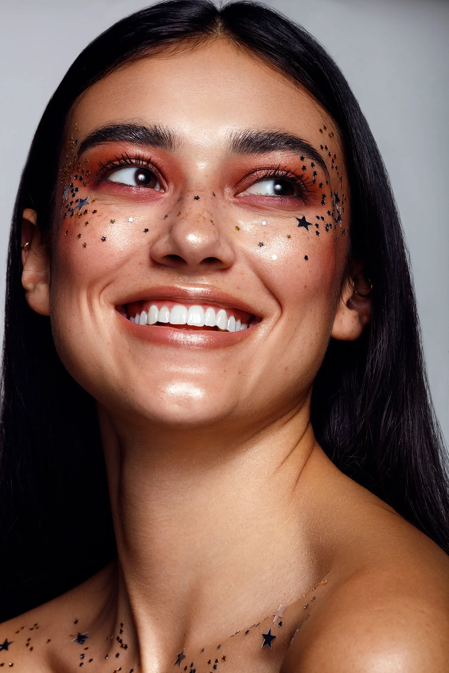 A woman with long dark hair smiling, decorated with black, gold, and silver star-shaped confetti on her face and shoulders, wearing makeup with warm tones. Beauty Photographer Los Angeles.