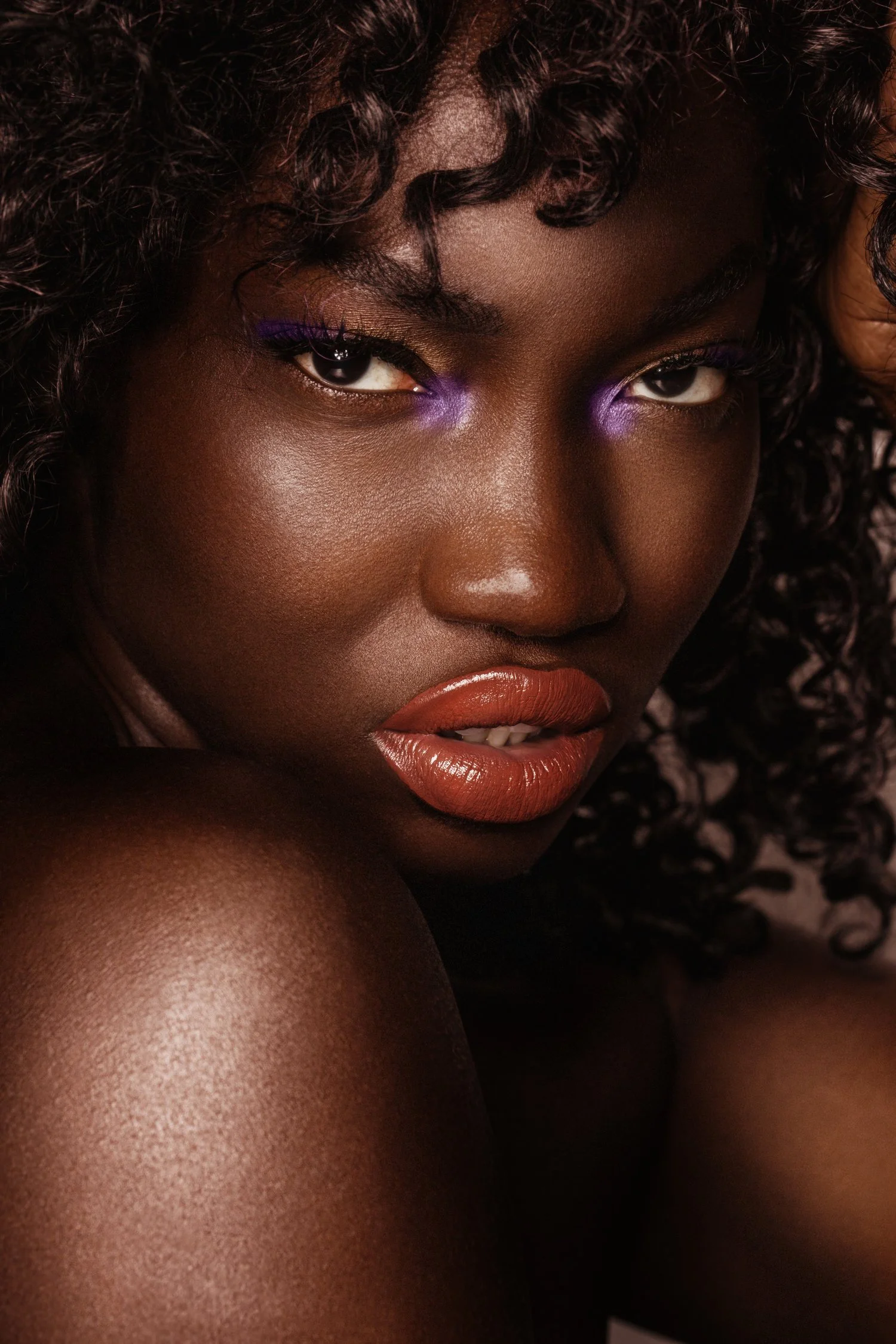 Close-up portrait of a woman with dark curly hair, wearing purple eye makeup, glossy lips, and smooth skin. Beauty Photographer Los Angeles.