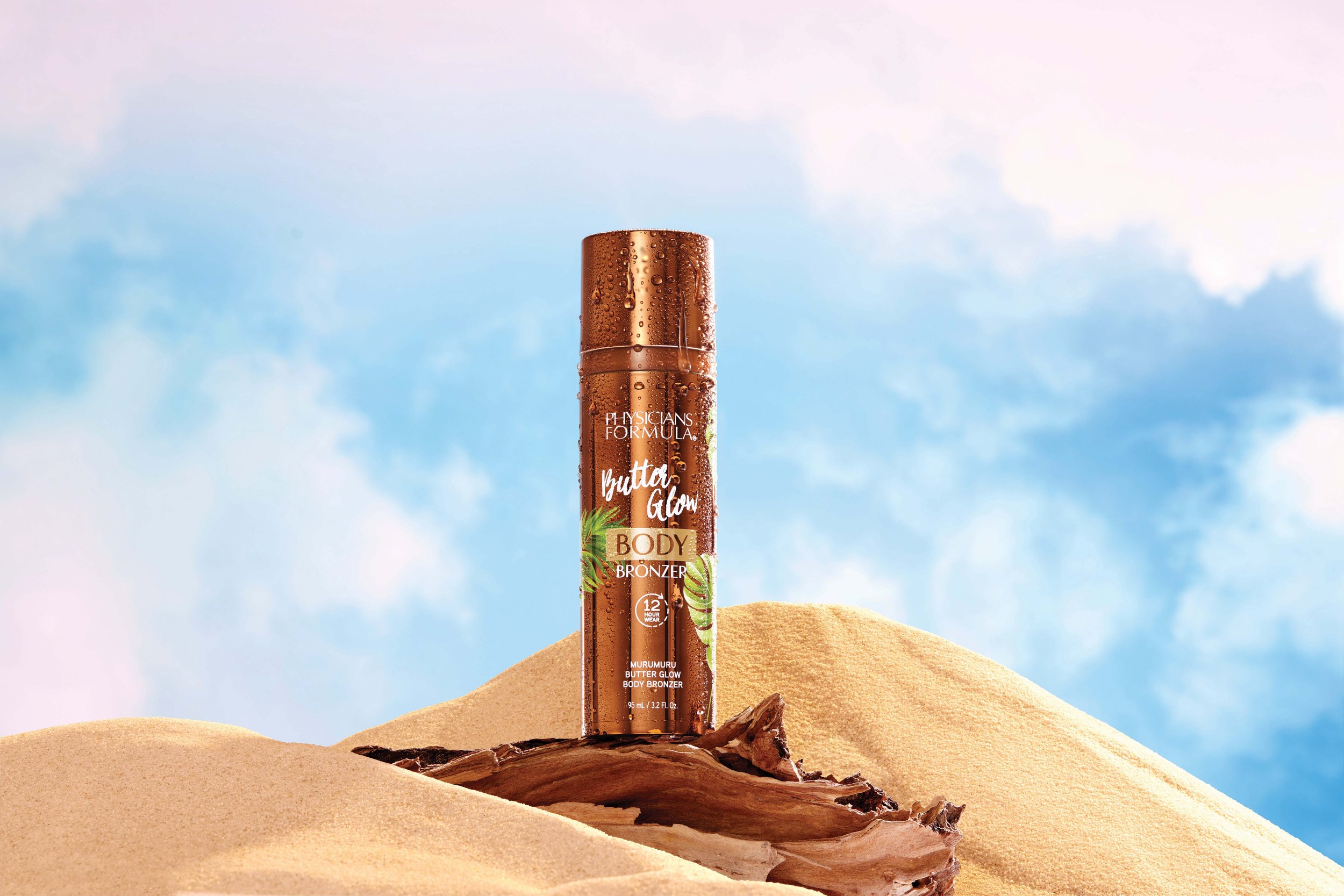 A can of Physcians Formula Butter Glow Body Bronzers is standing upright on a piece of driftwood on a sand dune with a blue sky and clouds in the background. Product photography Los Angeles.