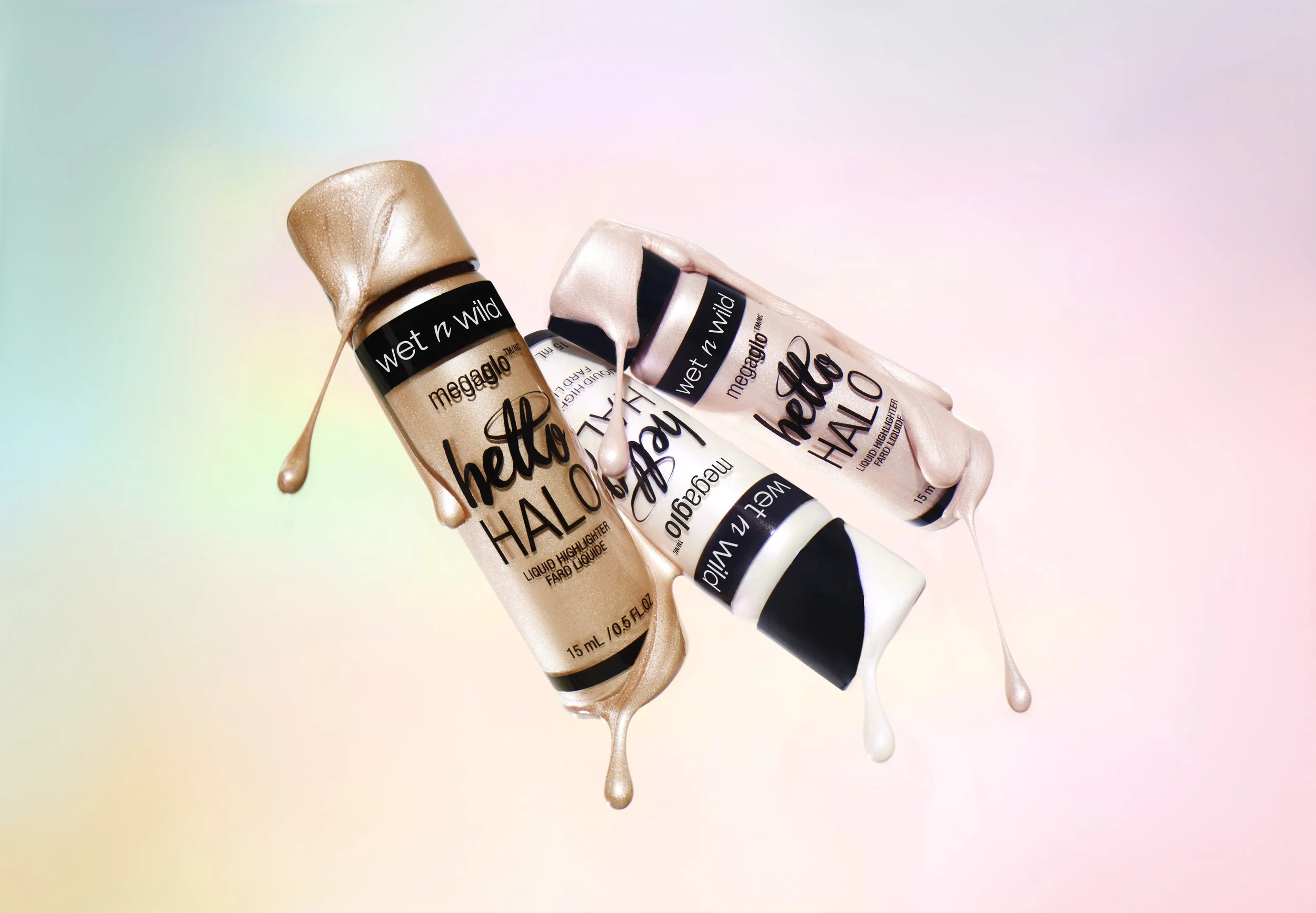 Three tubes of Wet n Wild MegaGlo Hello Halo liquid highlighter with beige, pink, and champagne shades, with their caps off and liquid highlighter drips on a pastel rainbow gradient background. Product photography Los Angeles.