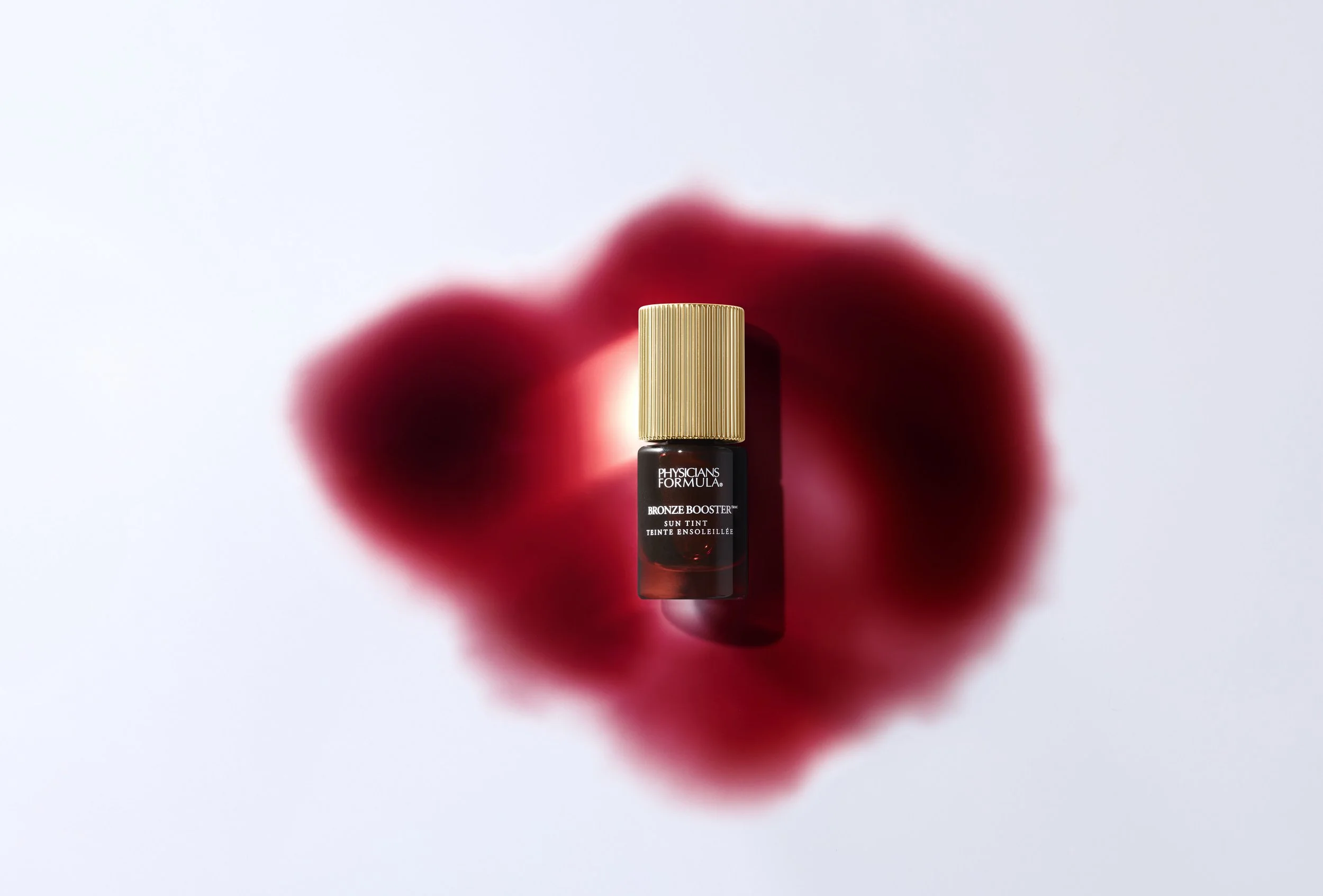 Brownen booster sun tint bottle with gold cap centered on white surface, shadow cast beneath, blurred red rose in the background. Product photography Los Angeles.