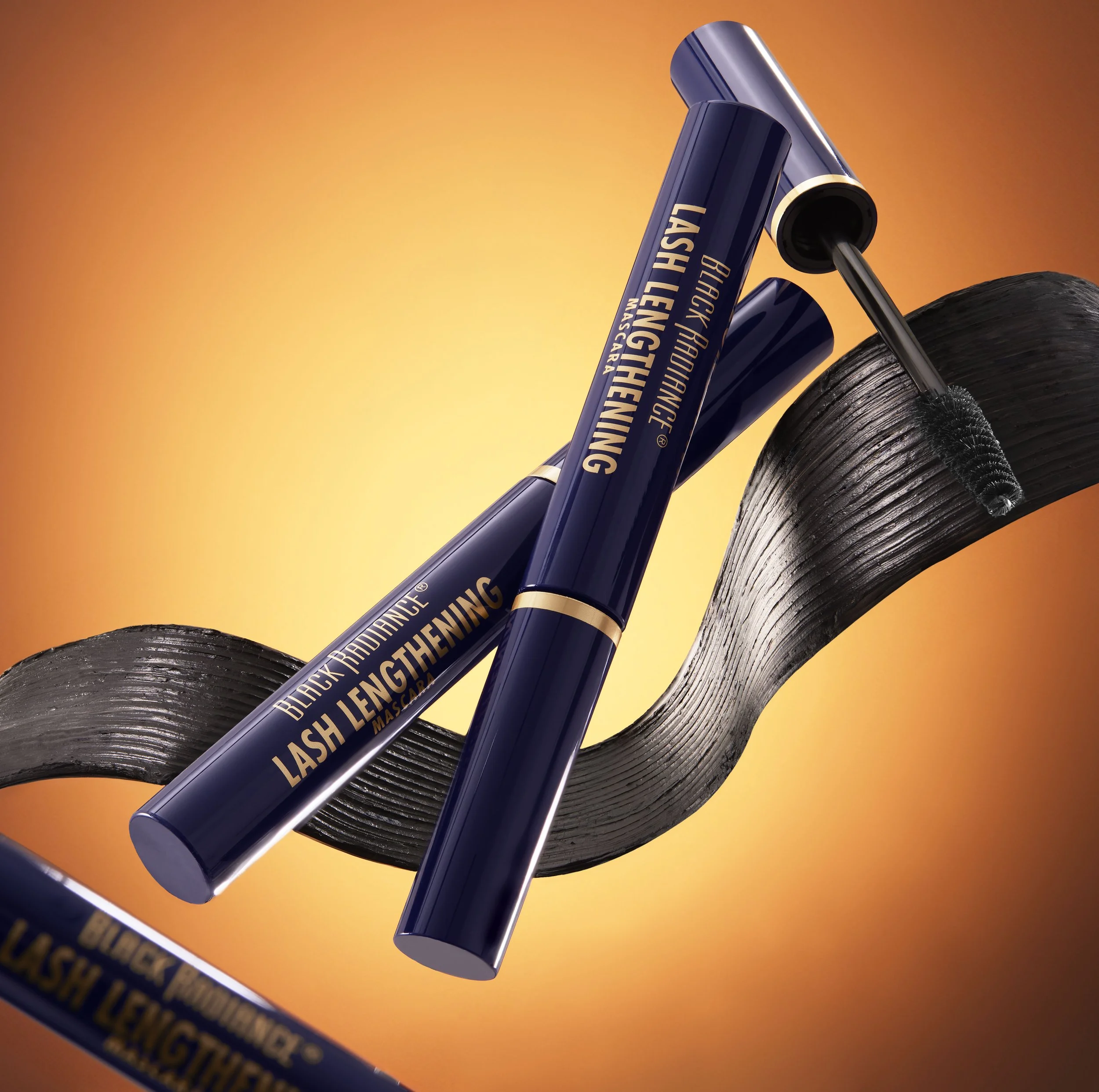 Two tubes of black mascara labeled 'Black Framing Lash Lengthening Mascara' resting on a shimmery black ribbon against a warm orange background. Product photography Los Angeles.