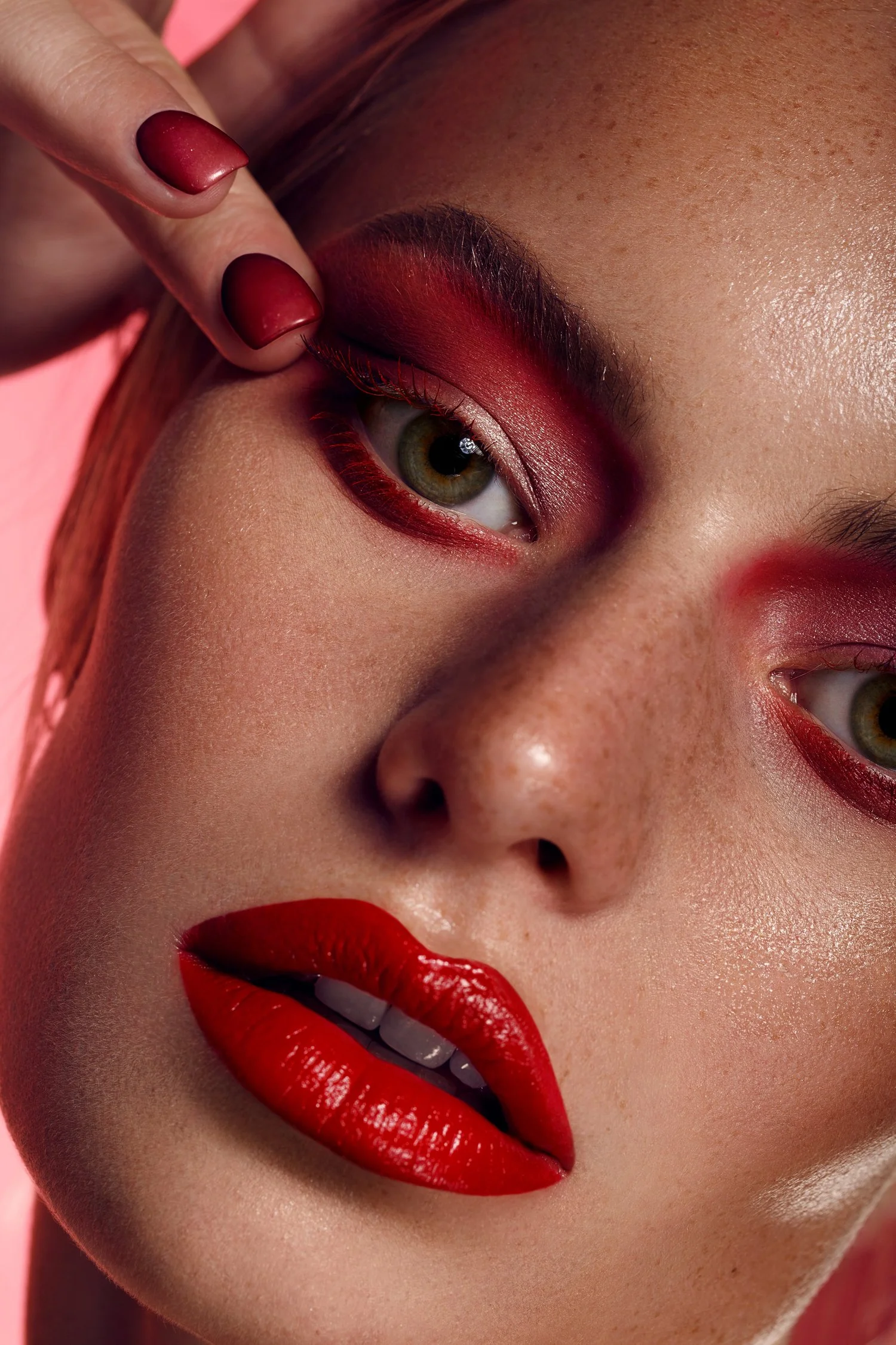 Close-up of a woman with bright red lipstick, red eyeshadow, and matching red nail polish. She is touching her eyelid with her index finger, displaying her makeup and facial features. Beauty Photographer Los Angeles.