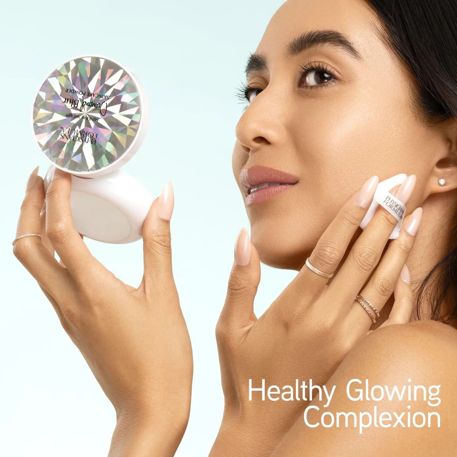 A woman with glowing skin applies a facial product using a spatula and holds a compact mirror. She has a light blue background and text that says 'Healthy Glowing Complexion.' Commercial Photographer Los Angeles.