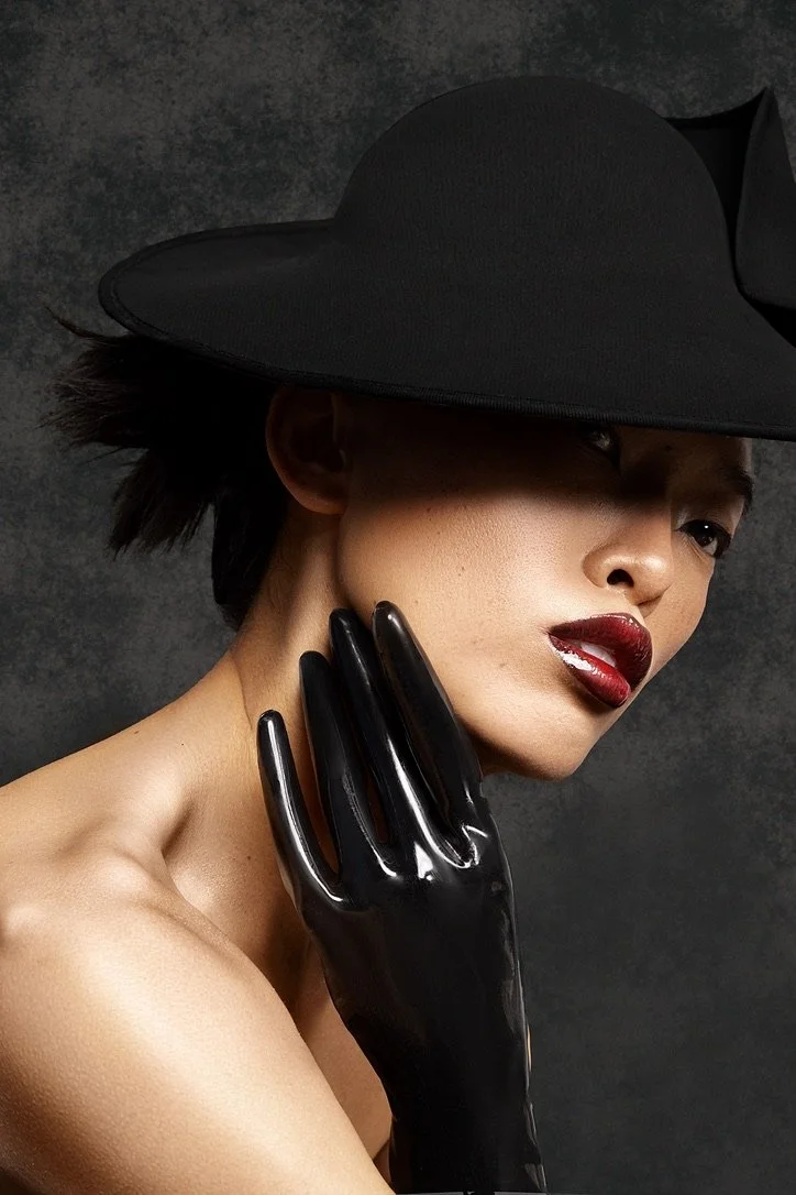 A woman with short dark hair wearing a large black hat and black gloves, with makeup including dark red lipstick, poses against a dark background. Beauty Photographer Los Angeles.