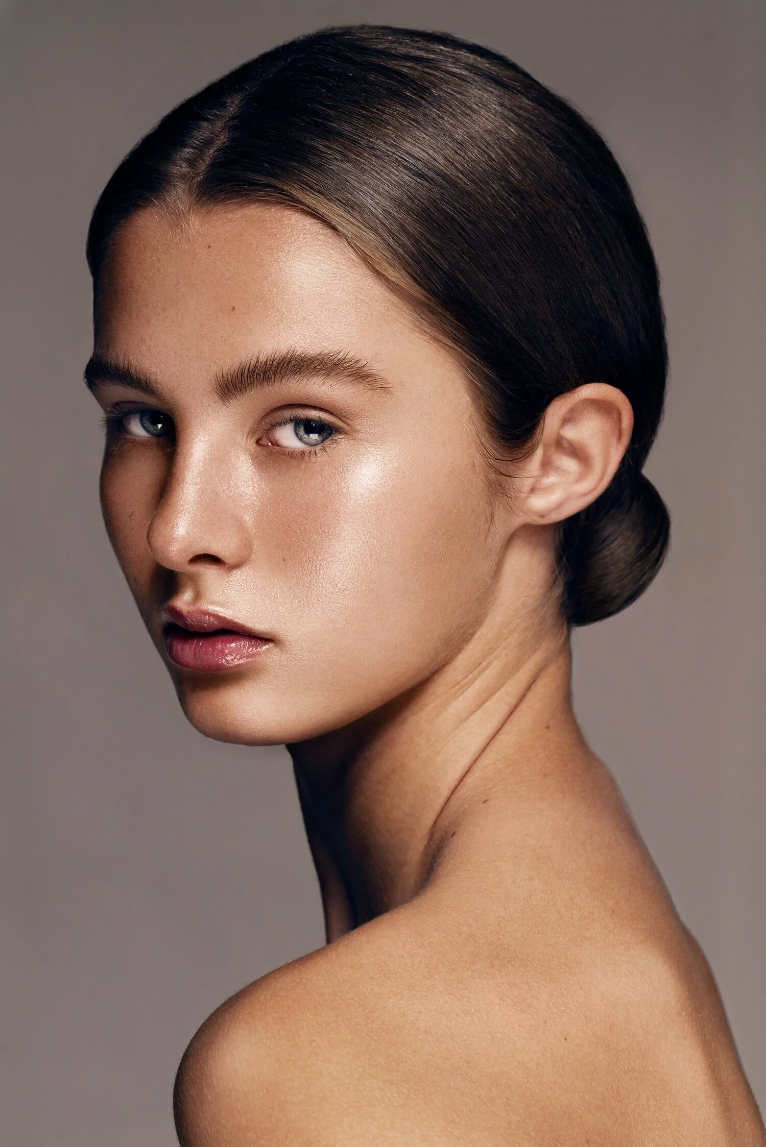 Close-up portrait of a young woman with smooth, shiny brown hair styled in a low bun, and clear skin, looking slightly to the left of the camera against a neutral background. Beauty Photographer Los Angeles.