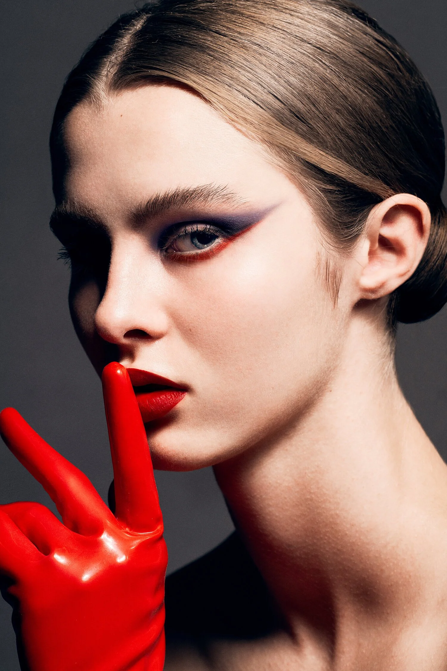 Close-up of a woman with dramatic makeup and short brown hair, wearing a red glove and red lipstick, with her finger held to her lips in a shushing gesture. Beauty Photographer Los Angeles.