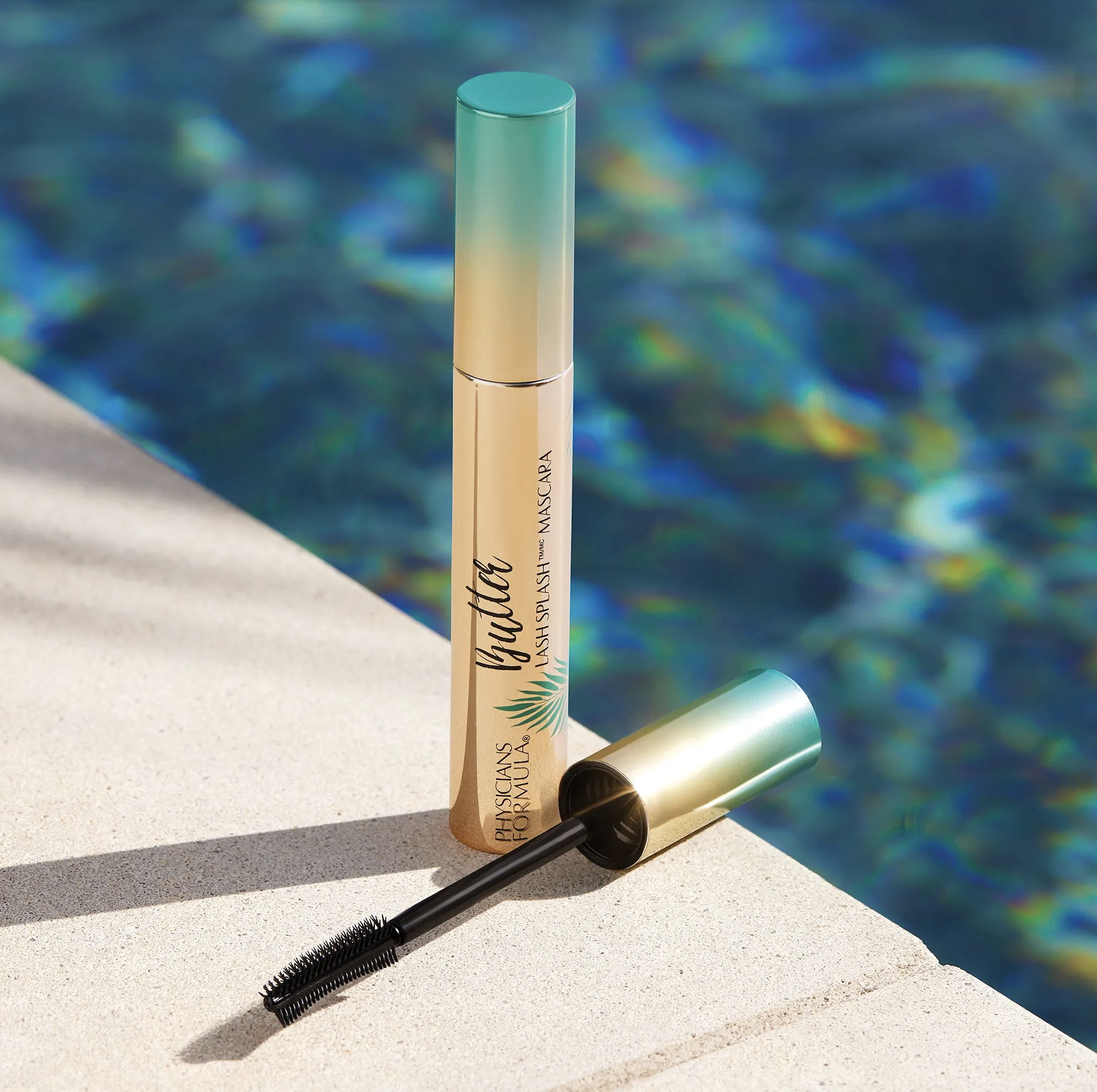A tube of mascara with a black wand lying on a poolside surface, with the tube standing upright. The tube is labeled 'Thirsty Lash Splash Mascara' and has a gradient blue and gold design. The background shows the blue water of a swimming pool. Produc