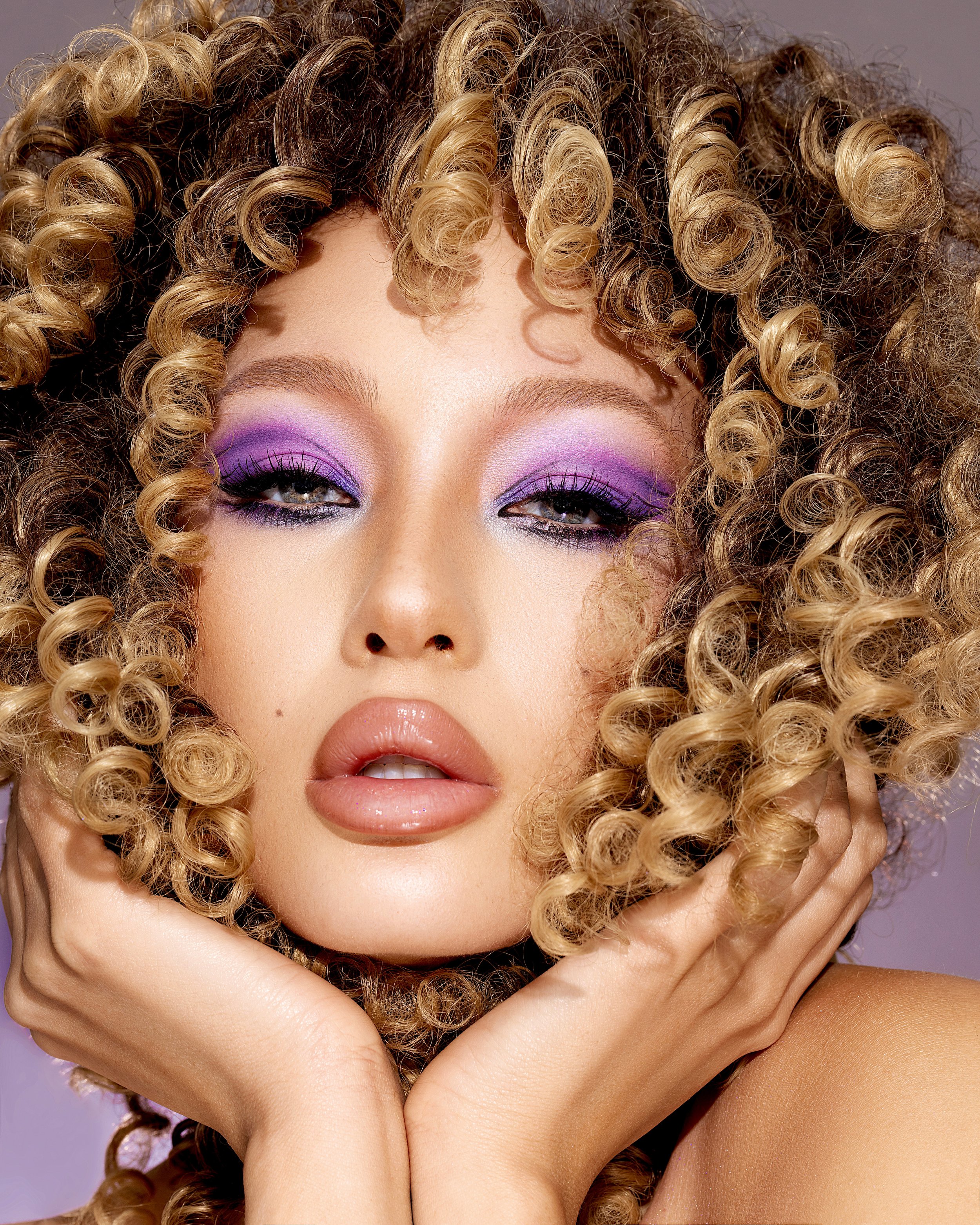 Close-up of a woman with voluminous curly blonde hair, purple eyeshadow, long eyelashes, and glossy lips, holding her face with both hands. Beauty Photographer Los Angeles.