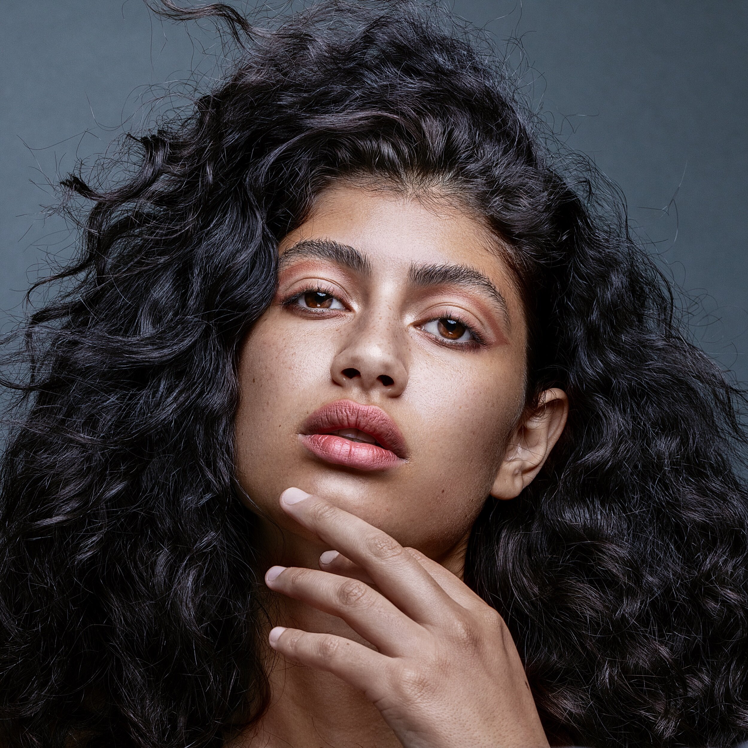 Close-up portrait of a woman with long, curly black hair, light skin, and brown eyes, touching her chin with her hand against a gray background.