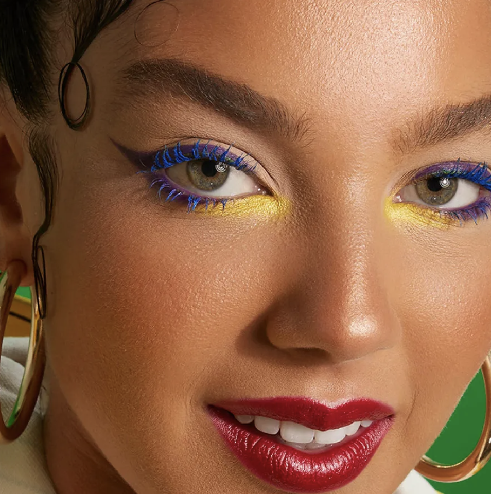 Close-up of a woman with colorful makeup, including yellow eyeshadow, purple eyeliner, and red lipstick, wearing large gold hoop earrings. Commercial Photographer Los Angeles.