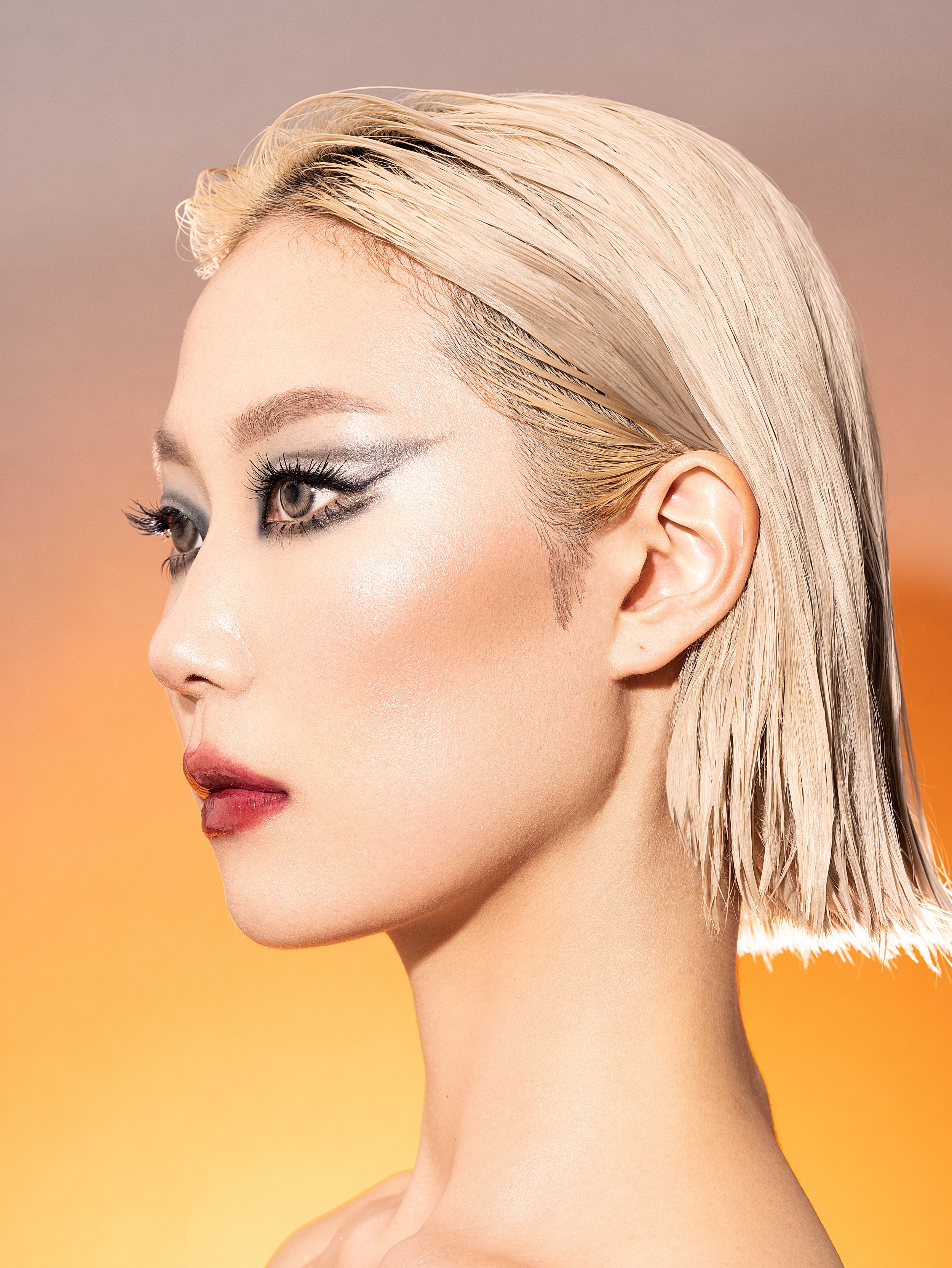 Close-up side portrait of a woman with platinum blonde wet hair, smoky eye makeup, and light skin against a warm gradient background. Beauty Photographer Los Angeles.