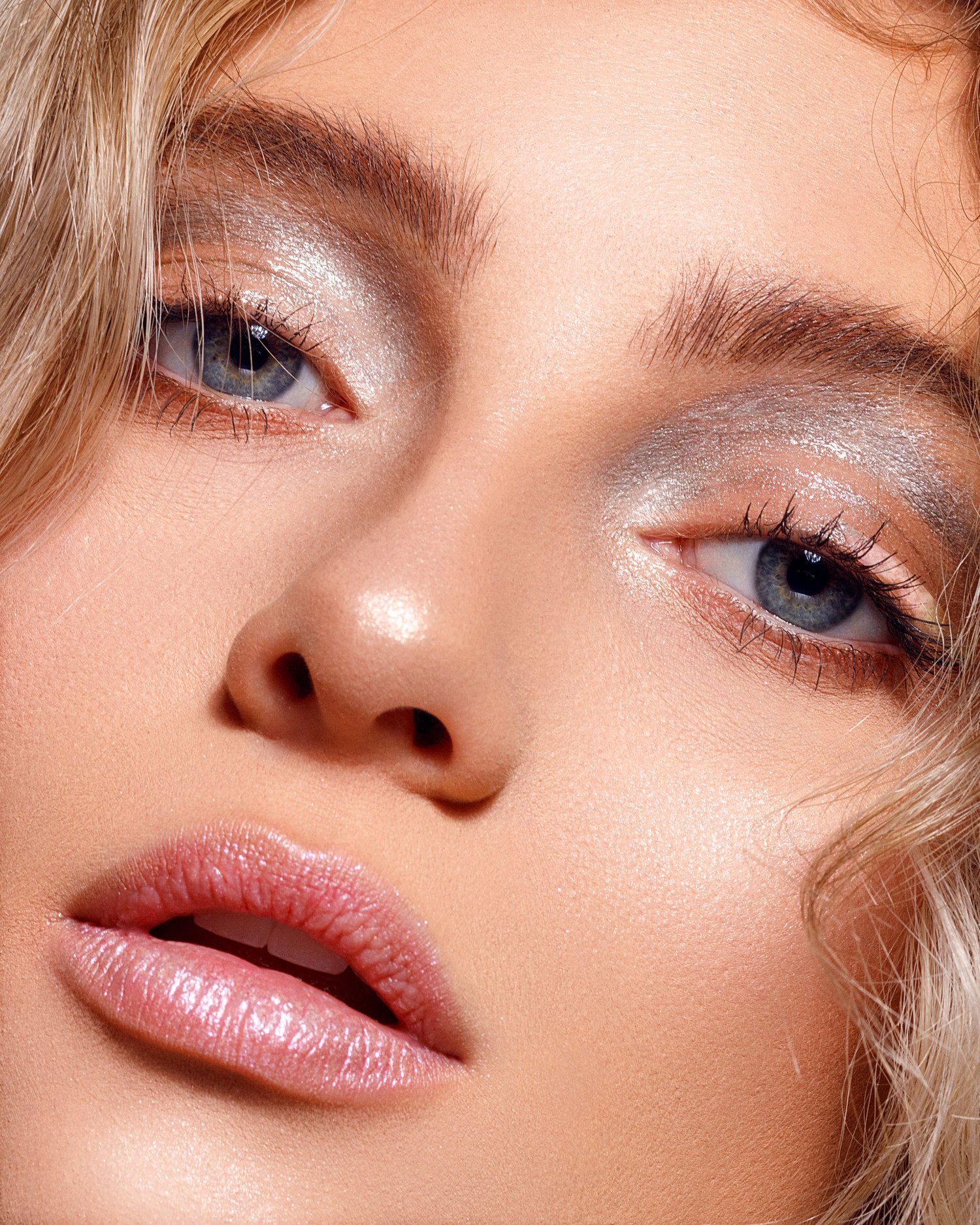 Close-up of a woman's face with blonde hair, showing her eyes, nose, and lips, with shiny makeup and smooth skin.