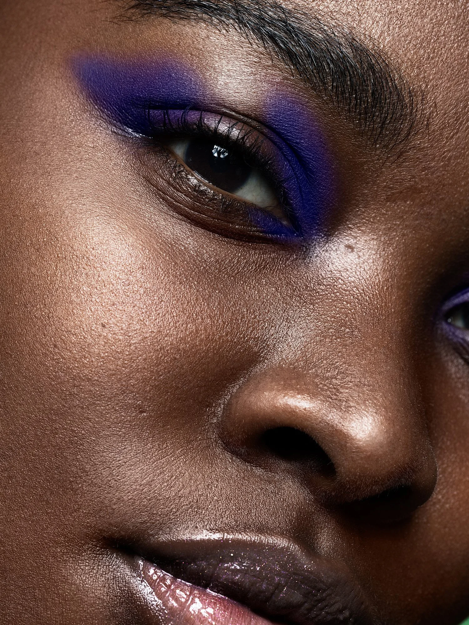 Close-up of a person with dark skin, focusing on the eye, nose, and lips, featuring bold purple eyeshadow and glossy lips. Beauty Photographer Los Angeles.