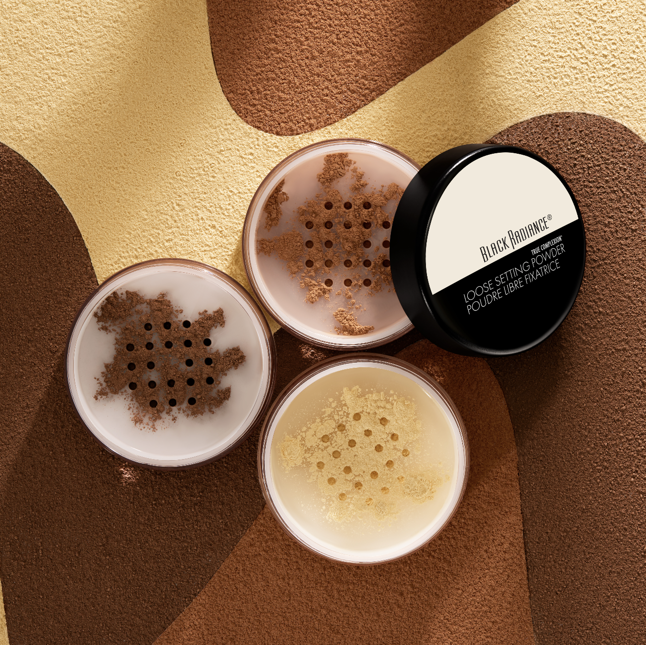 Three circular containers of loose setting powder in shades of brown, beige, and off-white, with one container's lid labeled 'Black Radiance Loose Setting Powder' on a textured, multicolored background. Product photography Los Angeles.