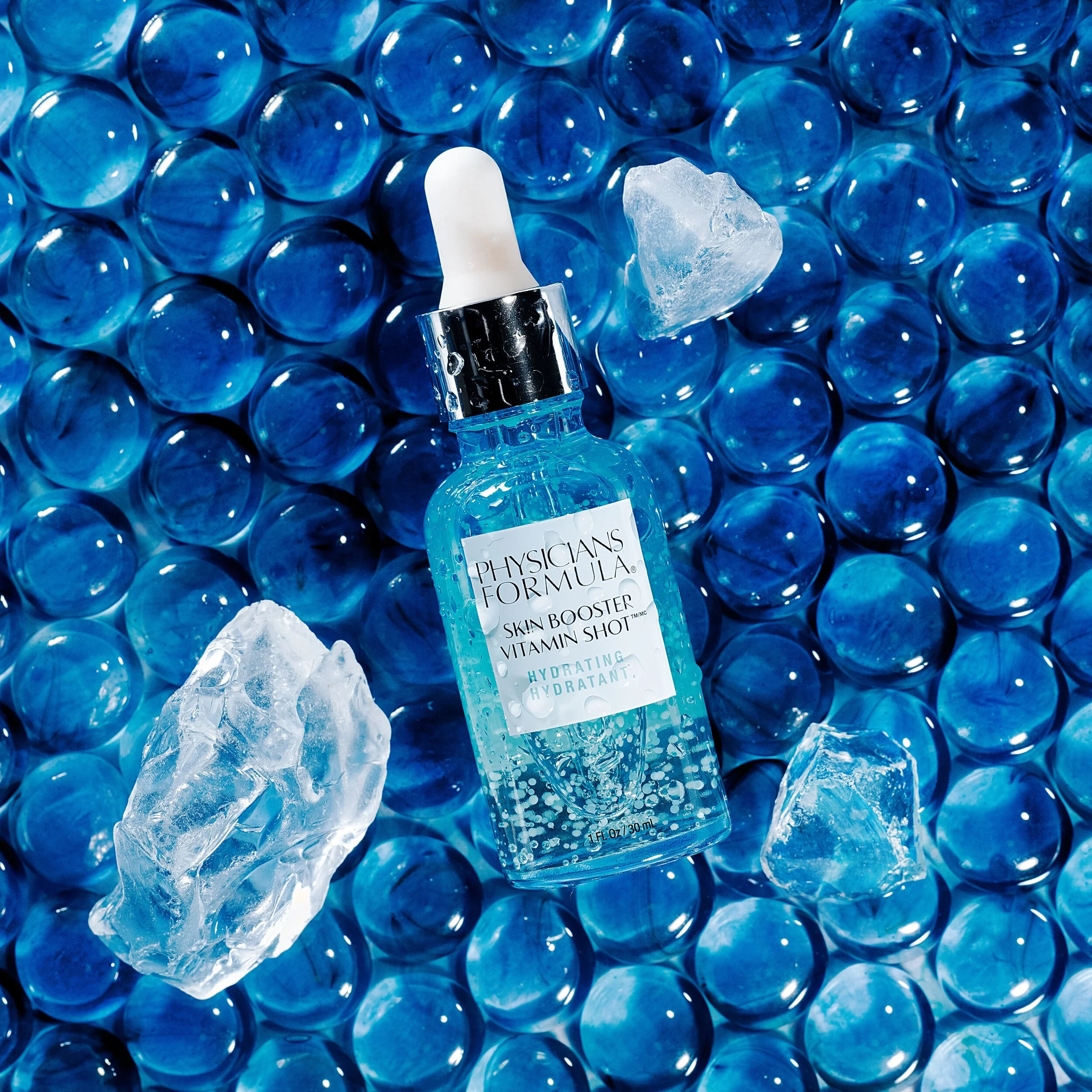 A bottle of Physicians Formula Skin Booster Vitamin Shot Hydrating serum surrounded by ice cubes on a background of blue bubbles. Product photography Los Angeles.