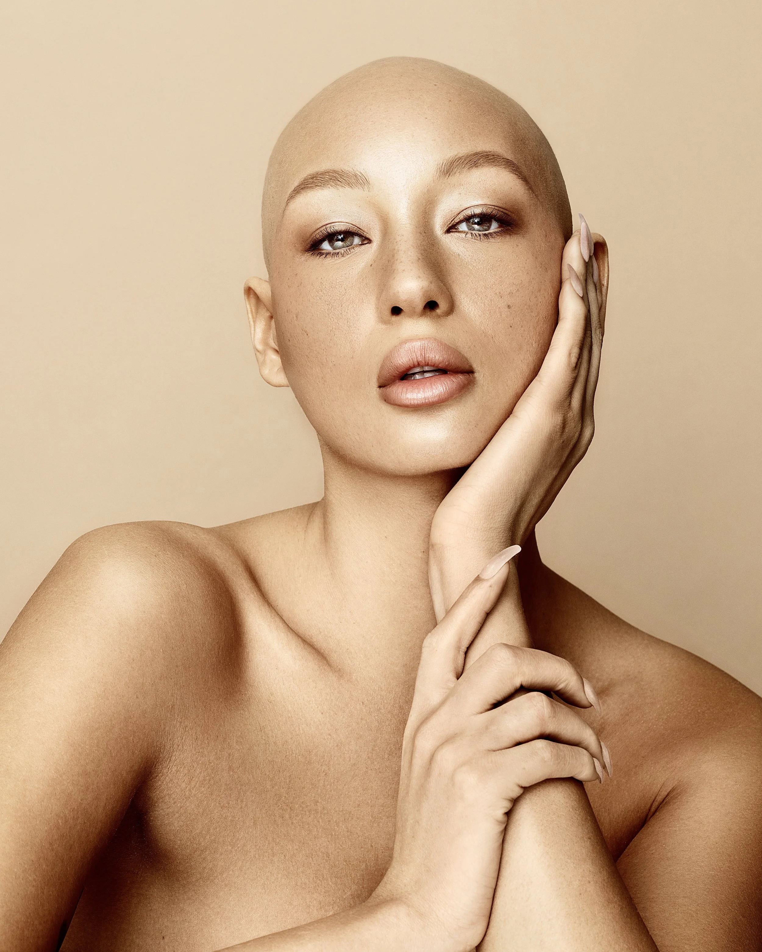Close-up of a woman with a shaved head, posing with her hand on her face against a beige background. Beauty Photographer Los Angeles.
