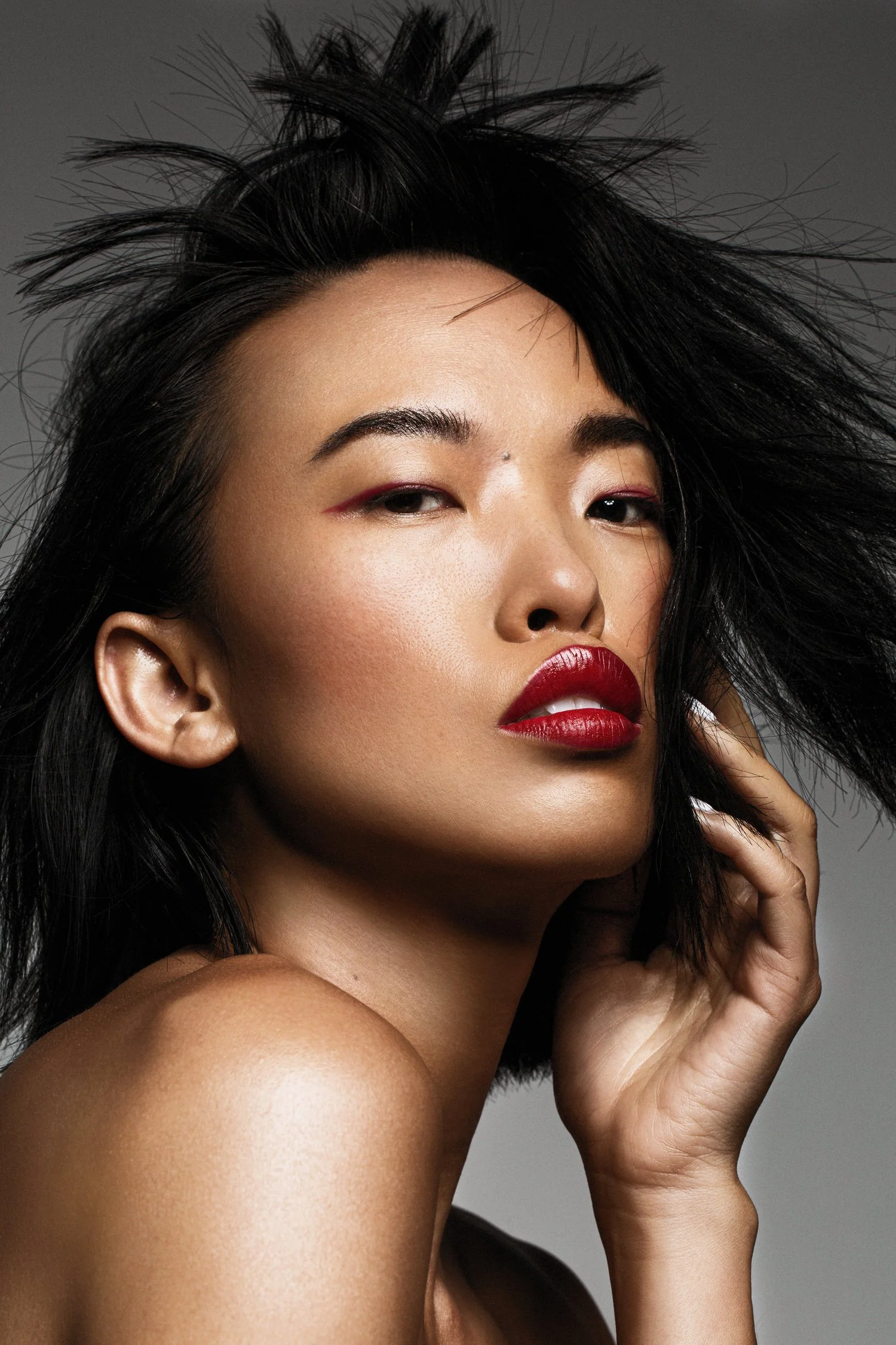 A woman with dark, tousled hair and bold red lipstick poses with one hand gently touching her face, against a gray background. Beauty Photographer Los Angeles.
