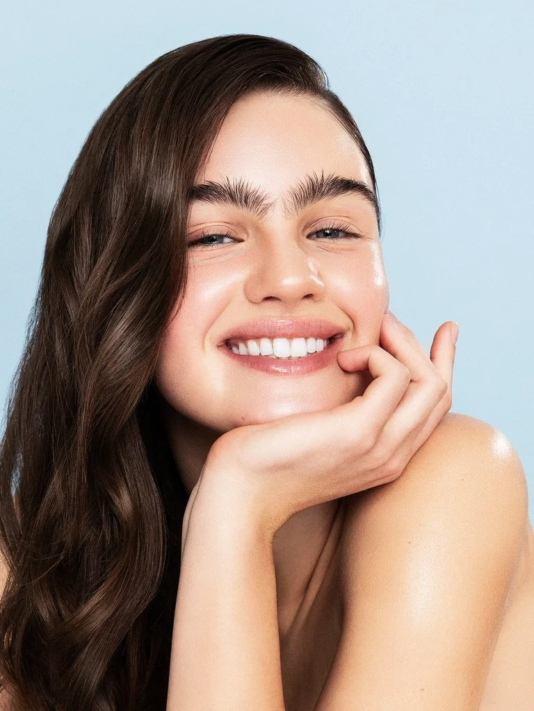 Close-up of a young woman with long, wavy brown hair smiling and resting her chin on her hand against a light blue background. Beauty Photographer Los Angeles.