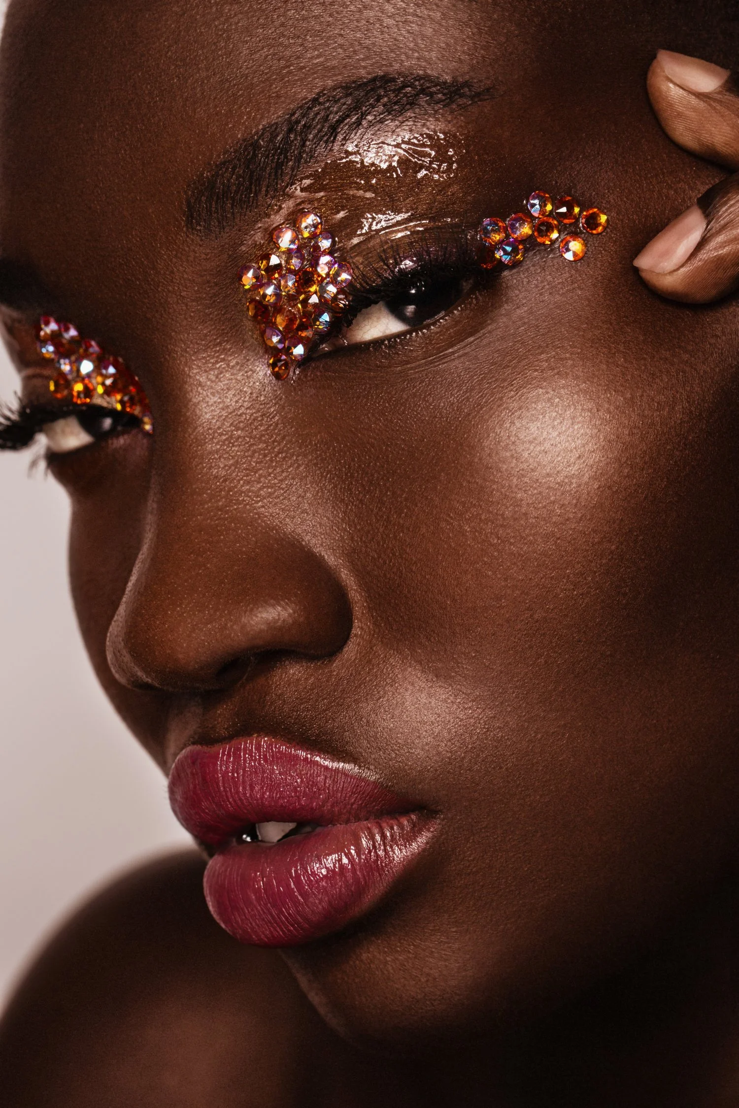 Close-up of a woman's face with dark skin, red lipstick, and rhinestones around her eyes, with one hand gently touching her temple. Beauty Photographer Los Angeles.