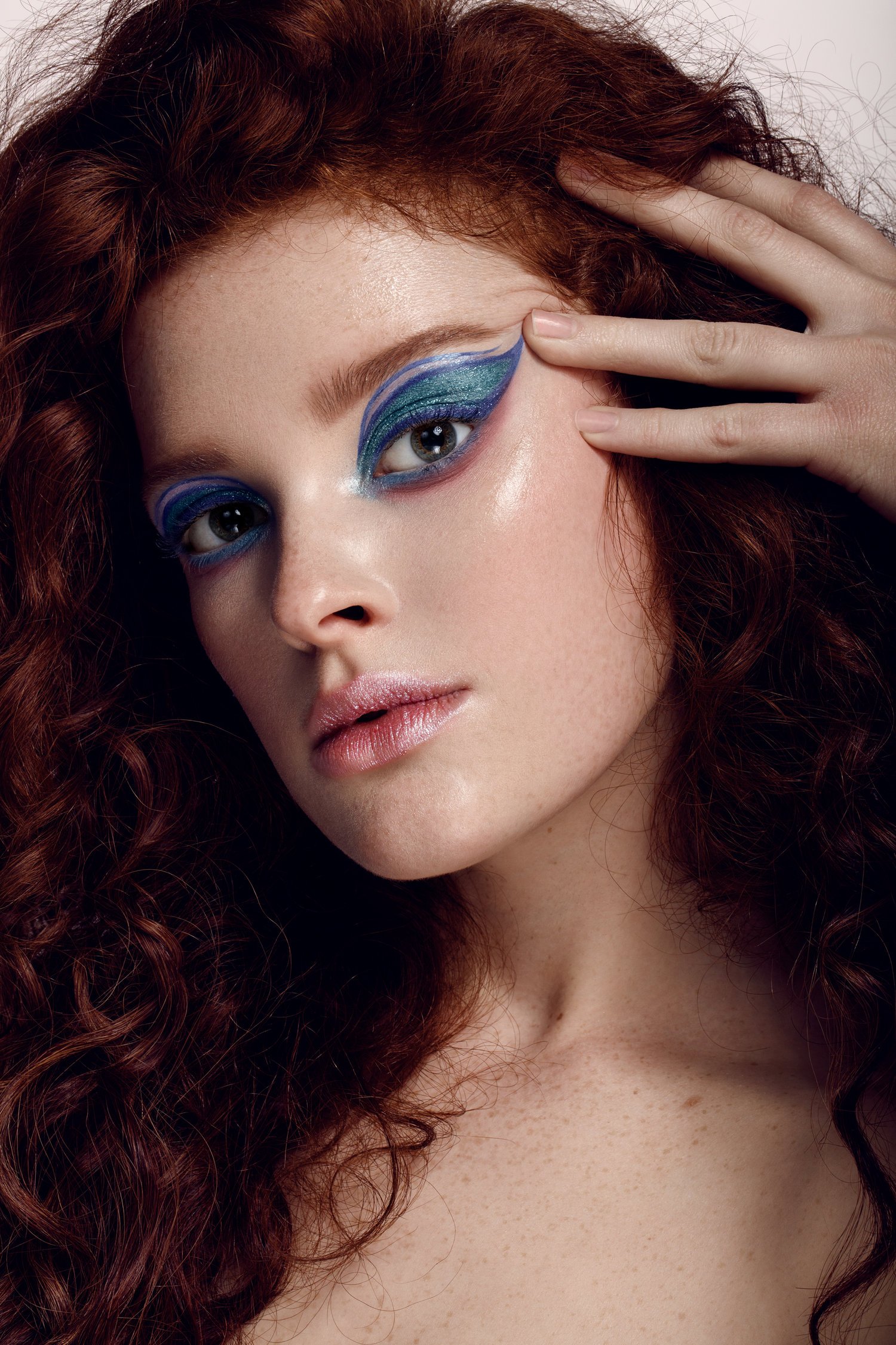 A woman with curly red hair and detailed blue eye makeup looks into the camera, touching her forehead with her right hand. Beauty Photographer Los Angeles.