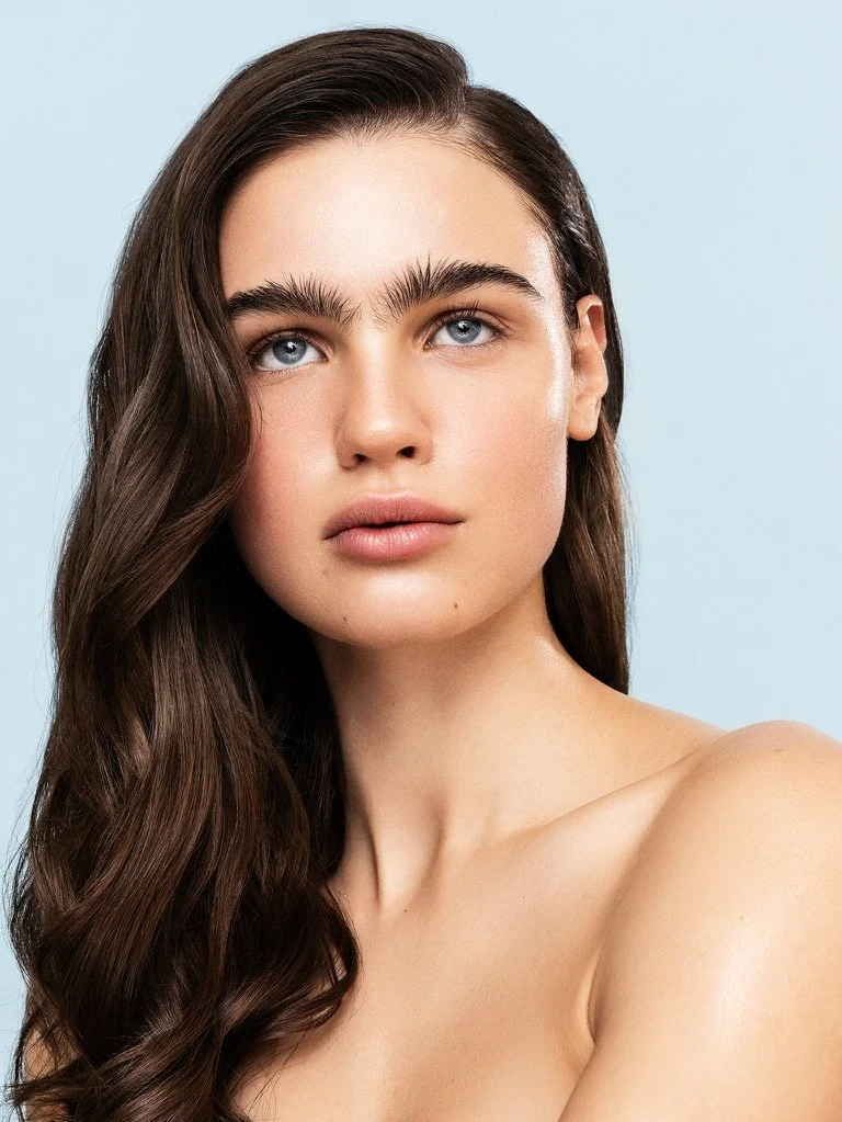 Close-up portrait of a young woman with long, wavy brown hair and piercing blue eyes against a light blue background. Beauty Photographer Los Angeles.