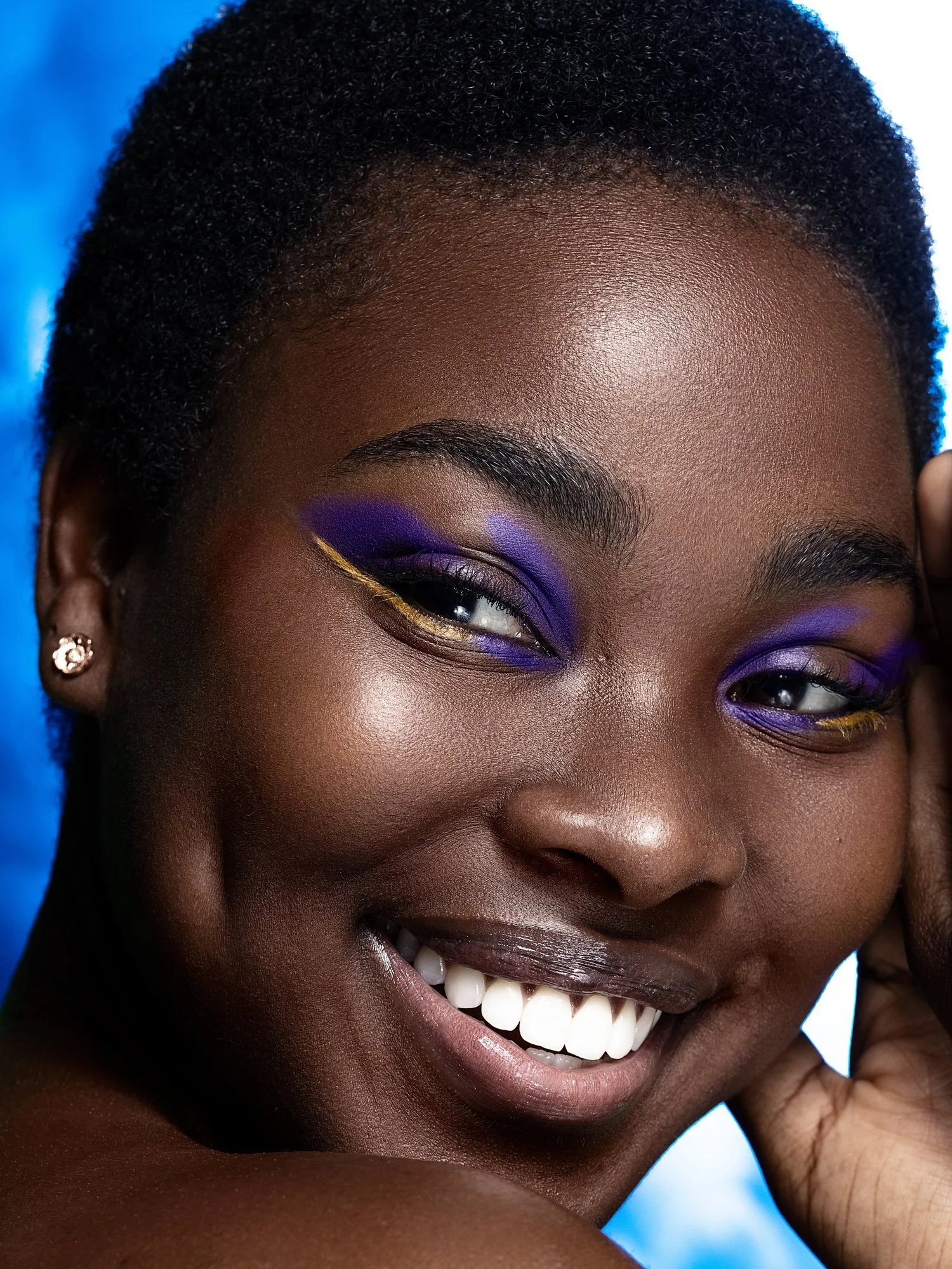 Close-up of a woman with short curly hair, vibrant purple and yellow eye makeup, smiling showing white teeth, wearing small earrings, against a blue background. Beauty Photographer Los Angeles.