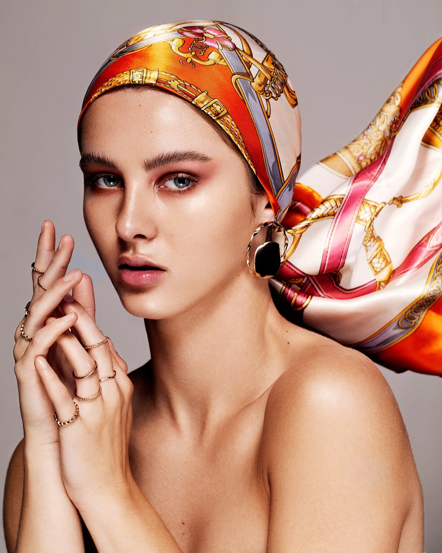 A woman wearing a colorful silk headscarf, large earrings, and multiple rings posing with her hands near her face against a neutral background. Beauty Photographer Los Angeles.
