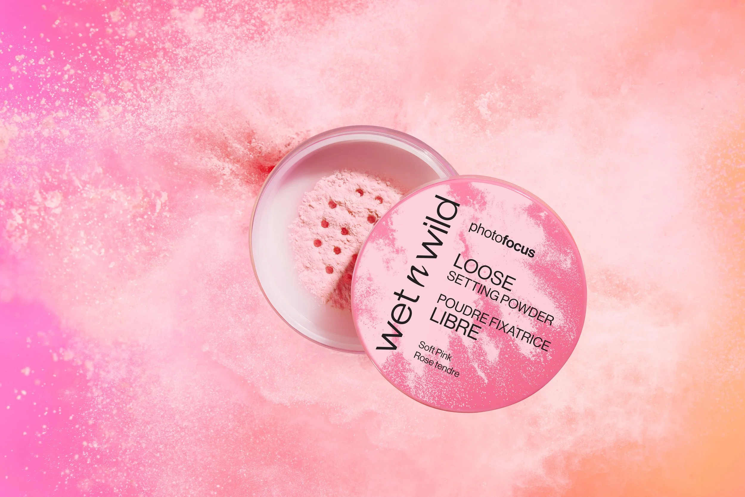 A round pink container of Photofocus loose setting powder with a partially open lid showing the pink powder inside, set against a pink and peach background. Product photography Los Angeles.