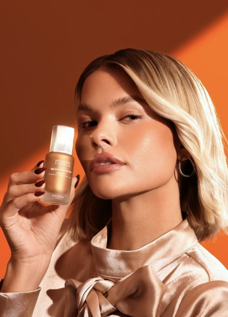 A woman with blonde hair holding a bottle of foundation near her face, posing against an orange background.
