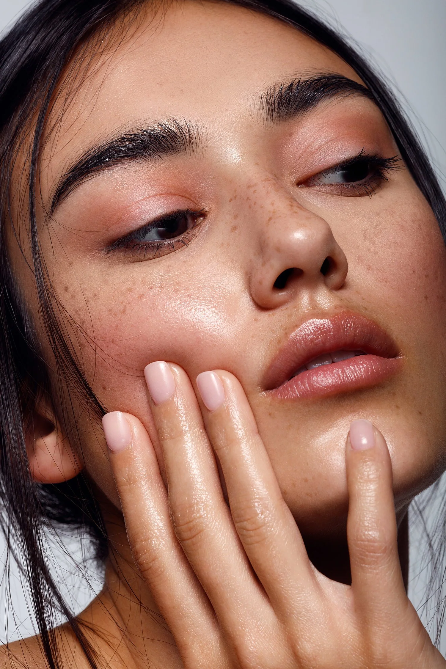 Close-up of a young woman's face with clear skin, freckles, and minimal makeup, touching her cheek gently. Beauty Photographer Los Angeles.