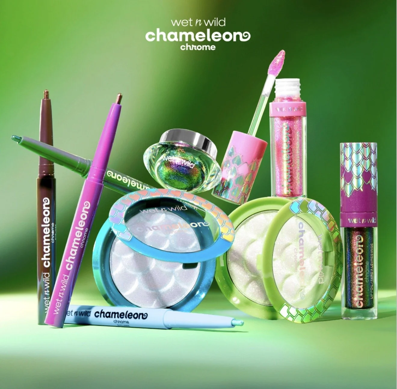 Collection of Wet n Wild Chameleon chrome cosmetics including lip gloss, powders, eyeliners, and a brush, with a green background.