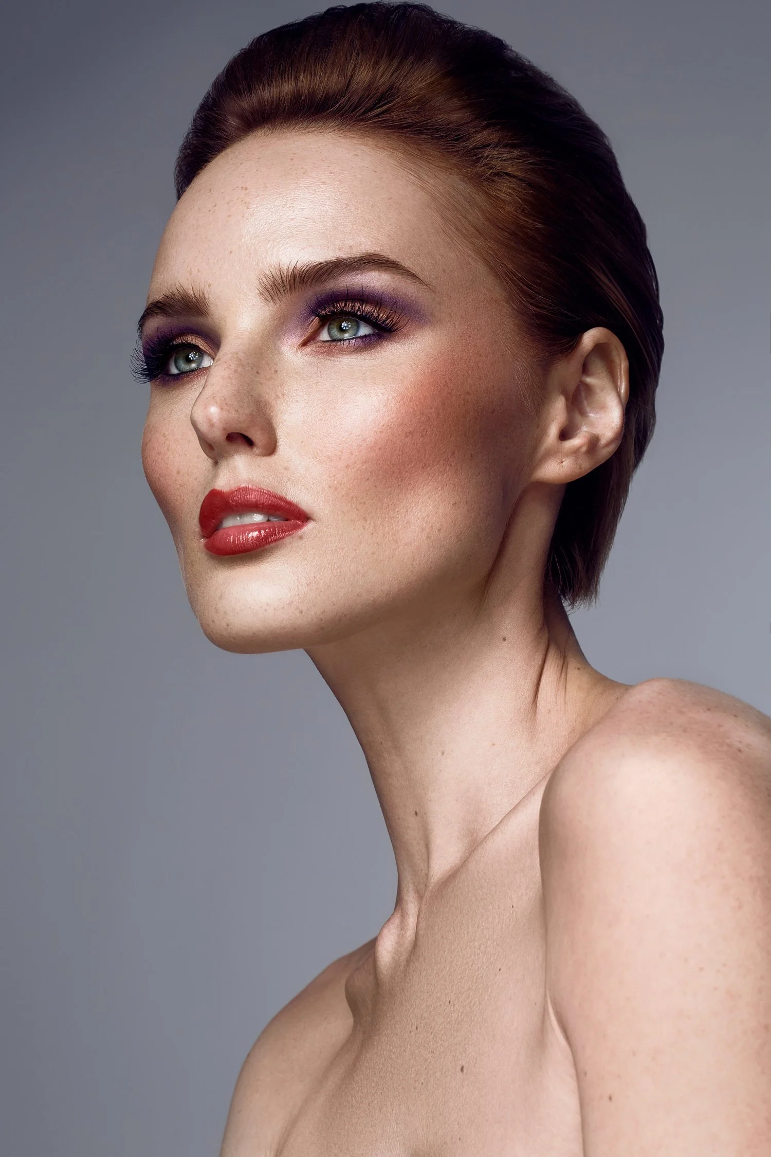 Close-up of a woman with red hair, fair skin, wearing makeup including purple eyeshadow, red lipstick, and long eyelashes, against a plain gray background. Beauty Photographer Los Angeles.