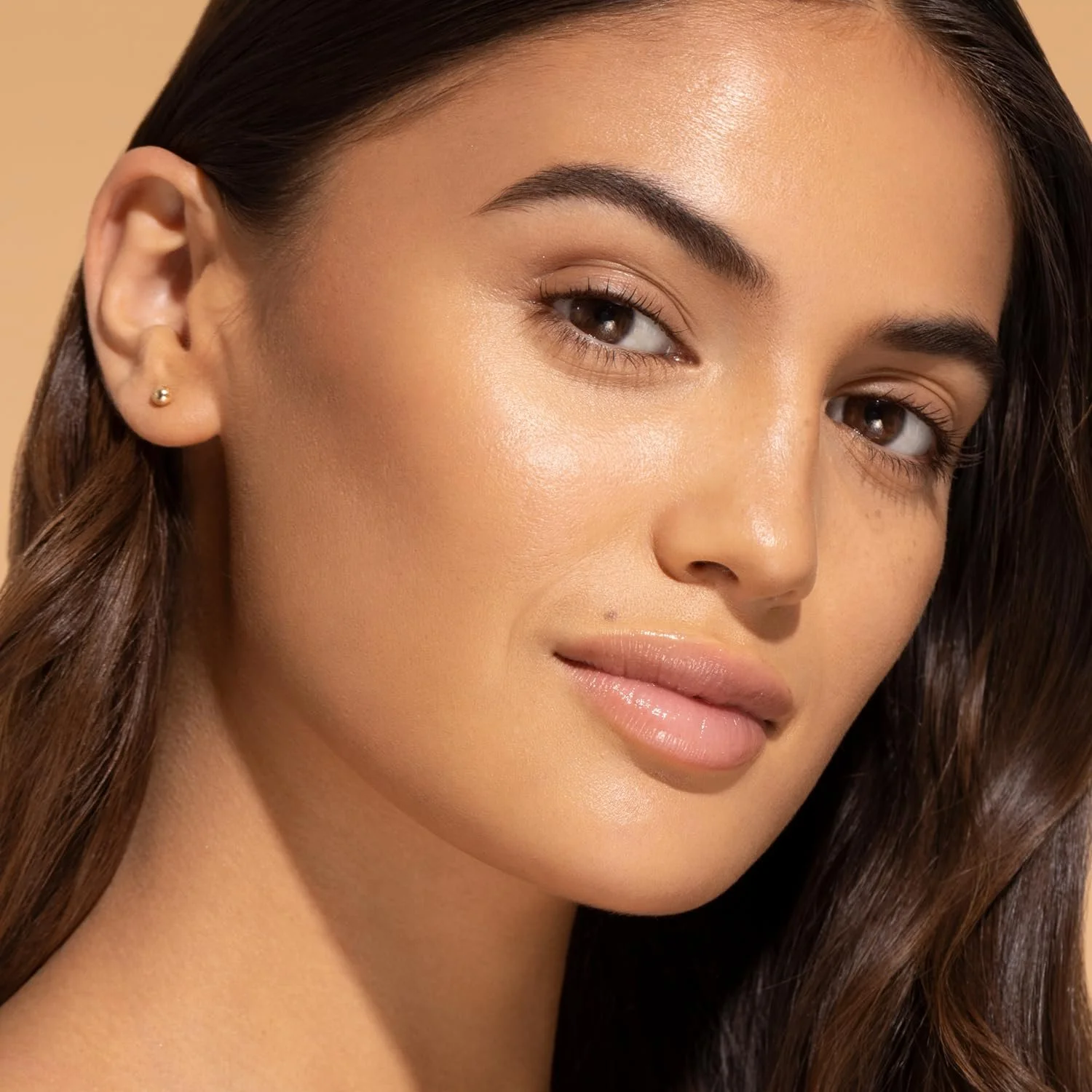 Close-up of a woman with smooth skin, brown eyes, and long brown hair, wearing small earrings, with beige background. Commercial Photographer Los Angeles.
