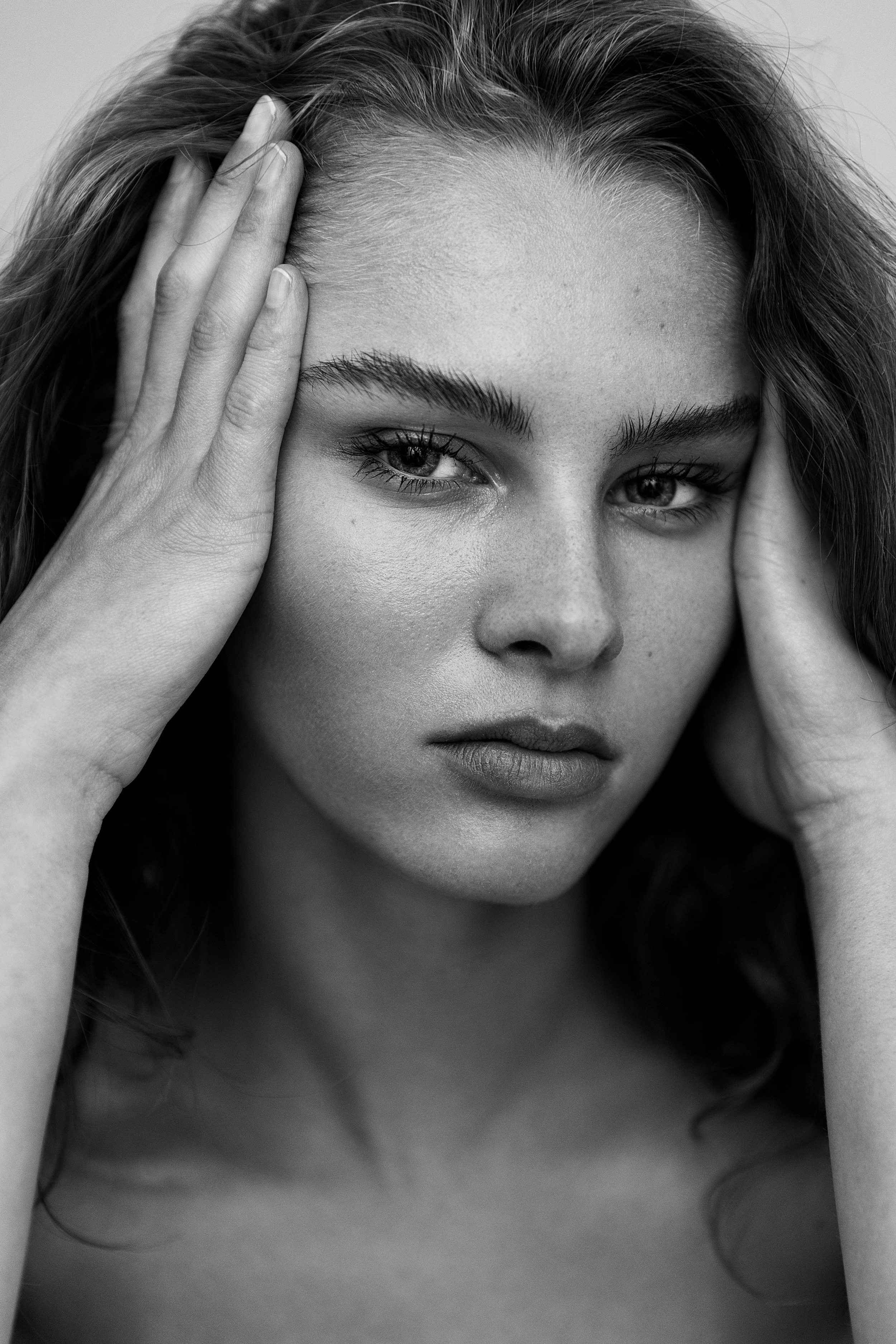 Black and white close-up portrait of a young woman with long wavy hair, holding her head with both hands, looking directly at the camera.