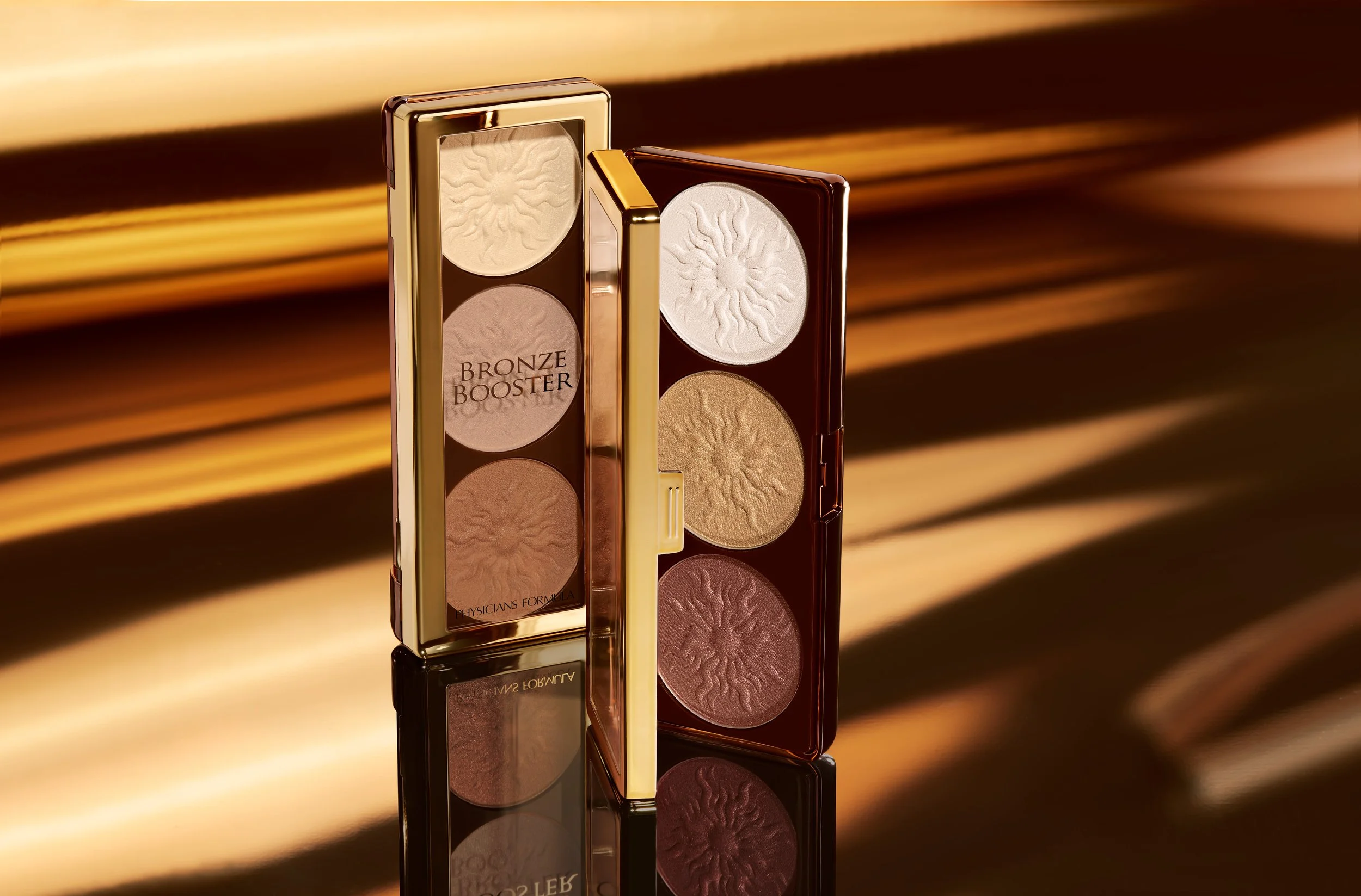 A makeup palette with three highlighter shades in a reflective gold and brown case, on a shiny surface with a golden striped background. Product photography Los Angeles.