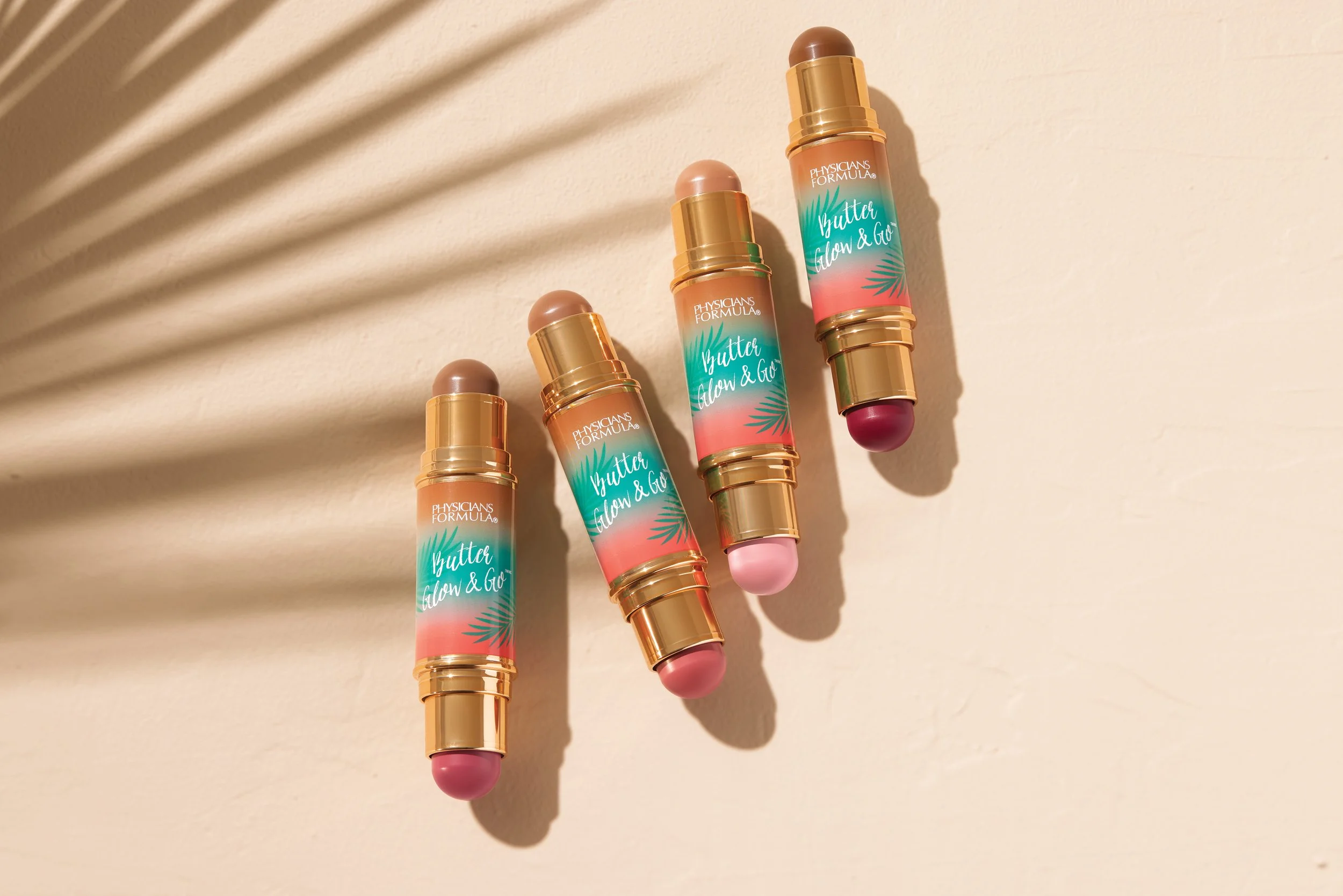 Four skincare sticks labeled 'Physicians Formula Butter Glow & Glo' with different shades, arranged on a beige wall, with shadows cast by a nearby plant. Product photography Los Angeles.