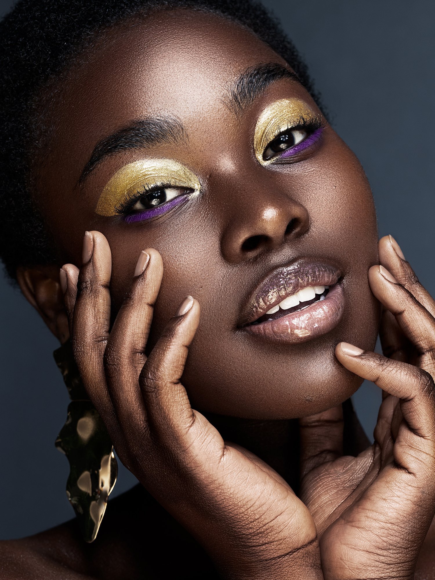 Close-up of a woman with dark skin, gold and purple eyeshadow, glossy lips, and gold earrings, touching her face with both hands. Beauty Photographer Los Angeles.
