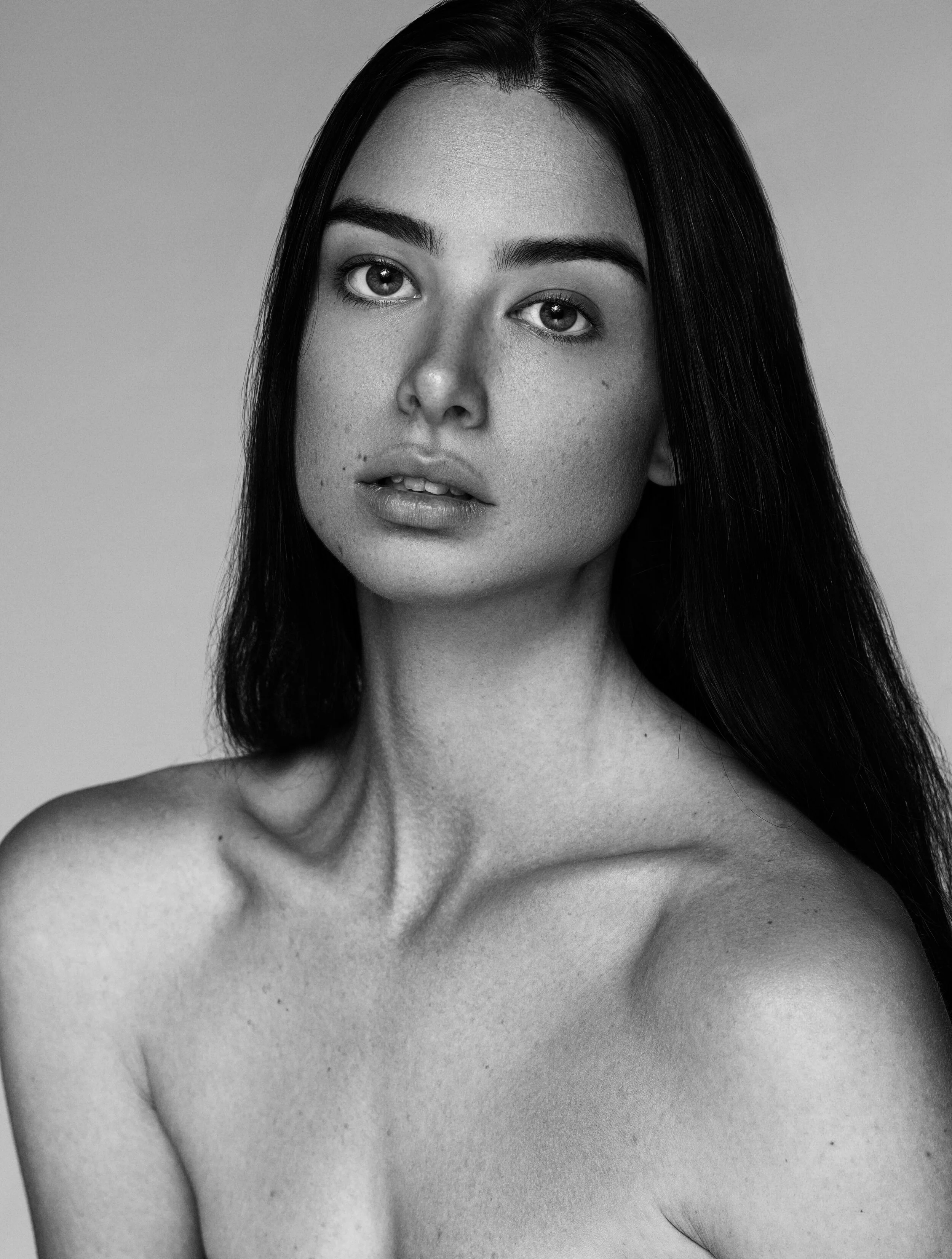 Black and white portrait of a young woman with long dark hair, looking directly at the camera with a neutral expression, without shirt, against a plain background.