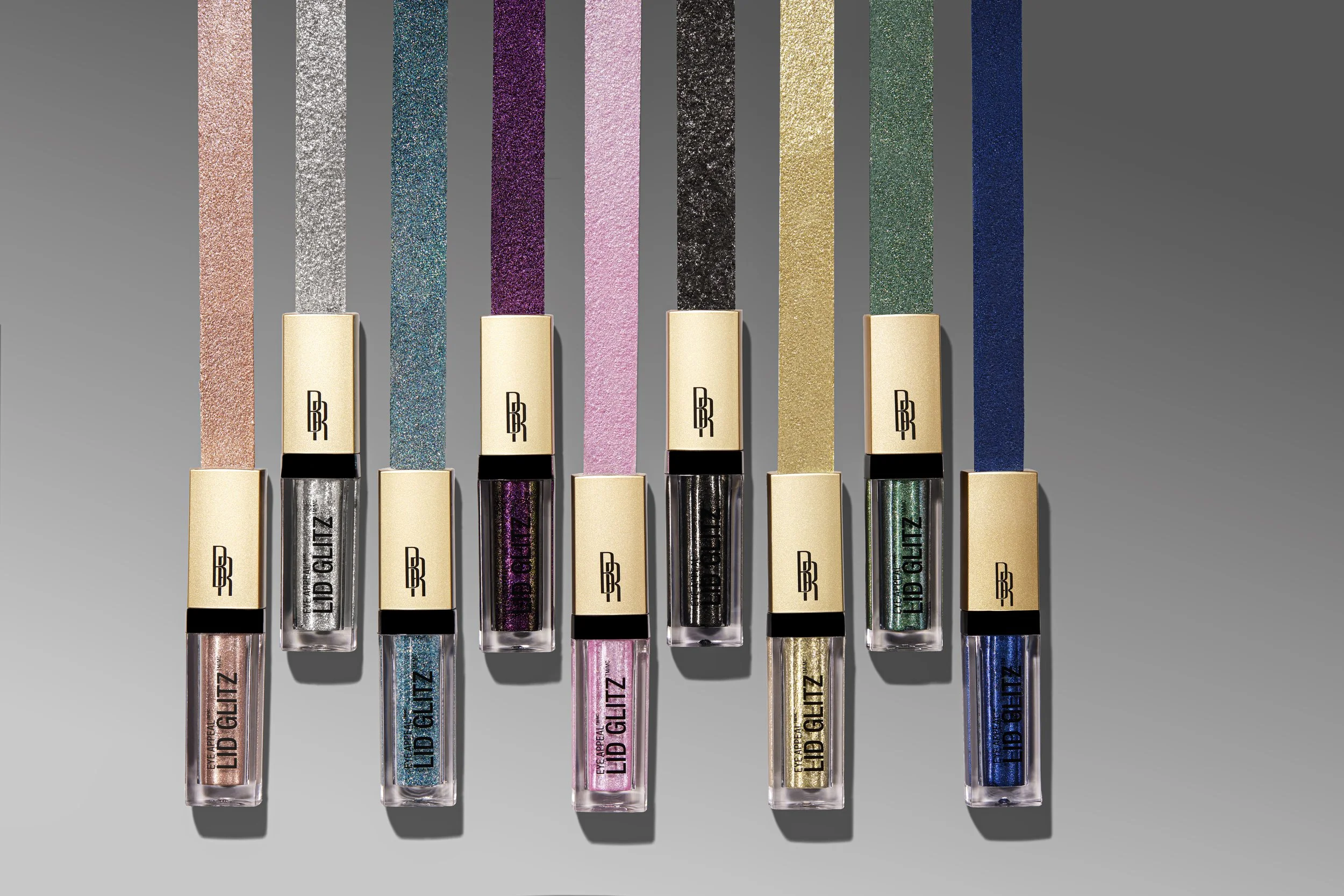 Set of seven lip gloss tubes with glittery, colorful caps in shades of pink, silver, purple, black, gold, green, and blue, arranged in a row on a gray background. Product photography Los Angeles.