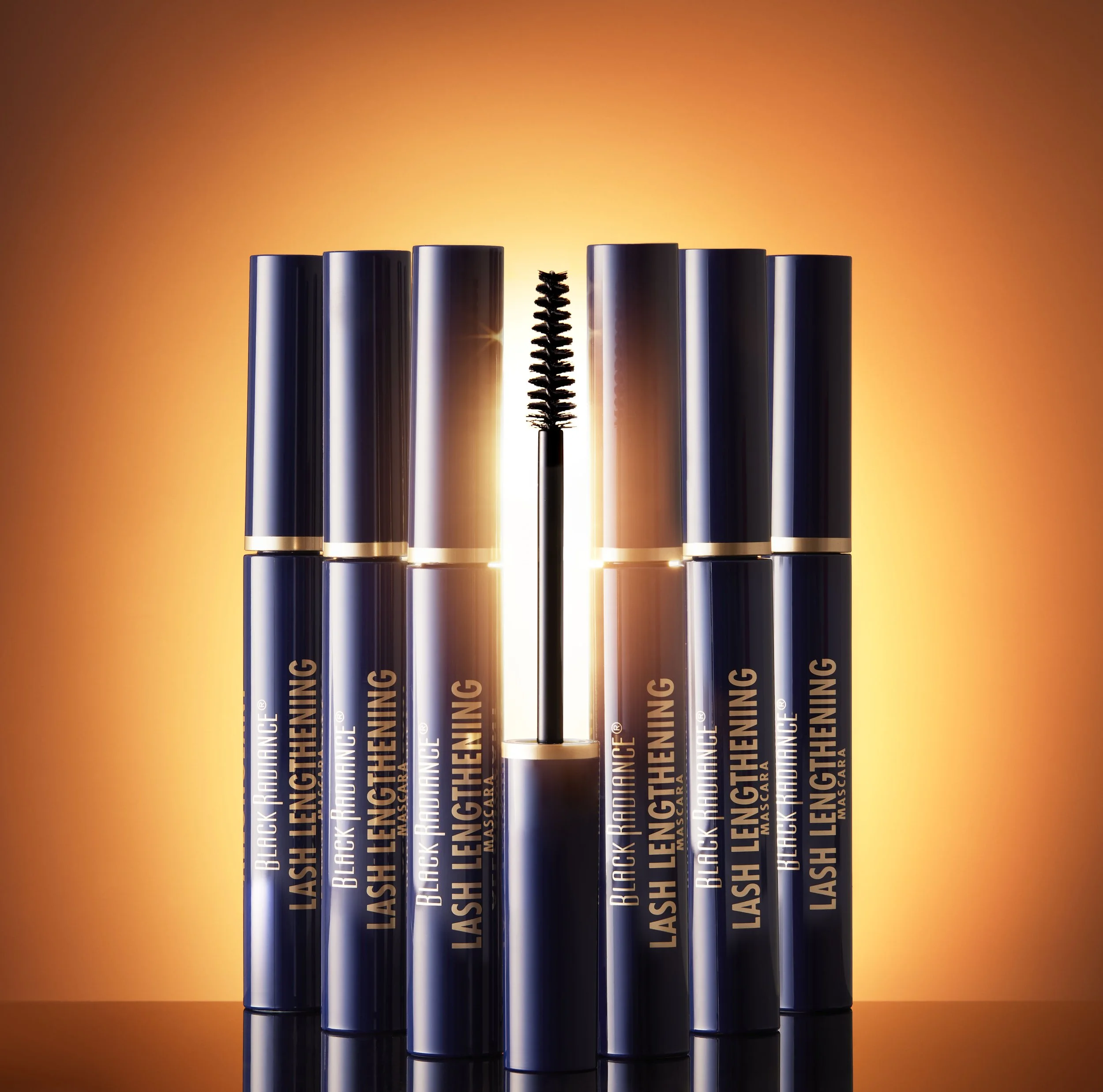 Six black mascara tubes with gold accents and one open mascara wand in the middle, against a warm glowing background. Product photography Los Angeles.