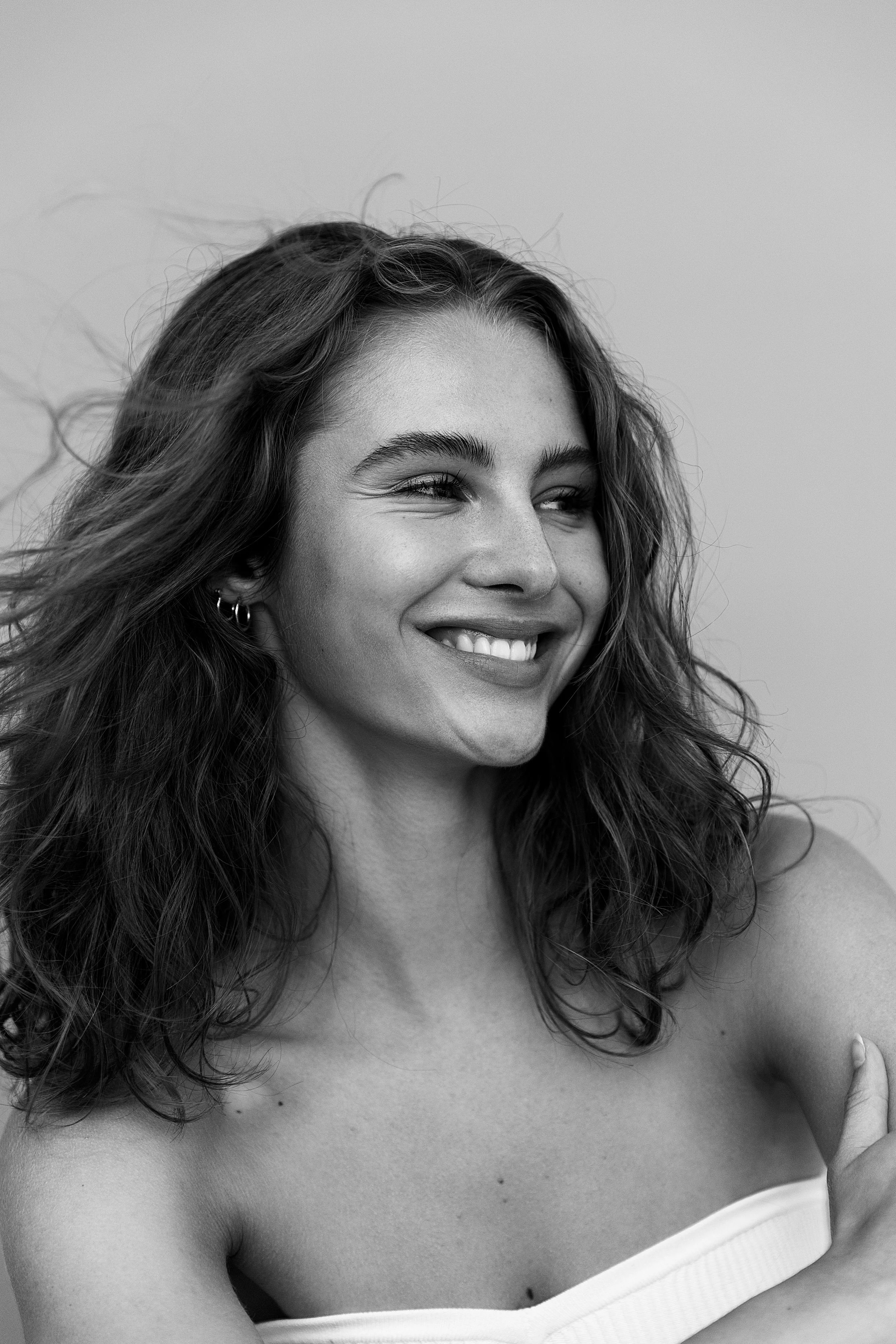 A smiling woman with wavy hair, wearing hoop earrings, in a strapless top, captured in black and white.