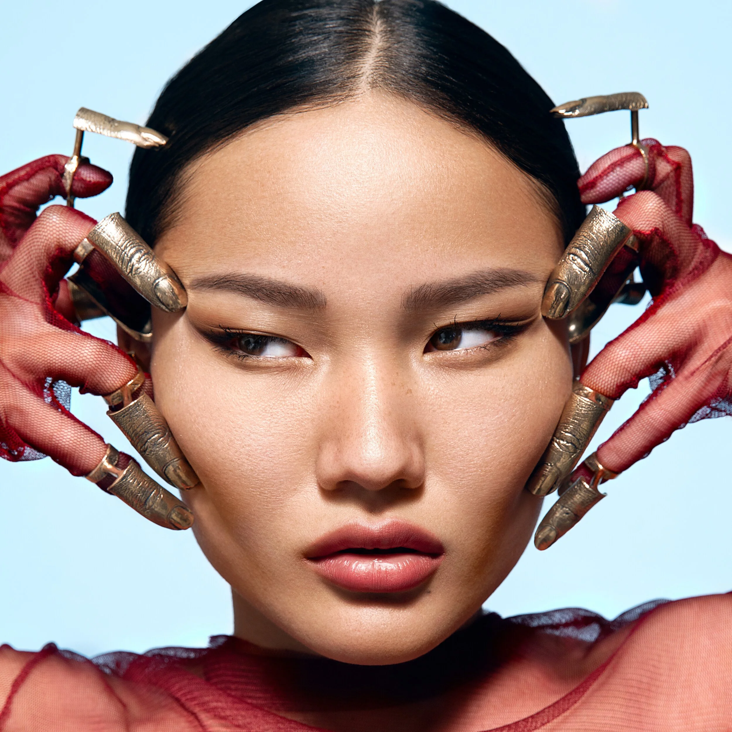 Close-up of a young woman with dark hair, makeup, and red mesh gloves, holding her head with metallic gloves around her face, against a light blue background.
