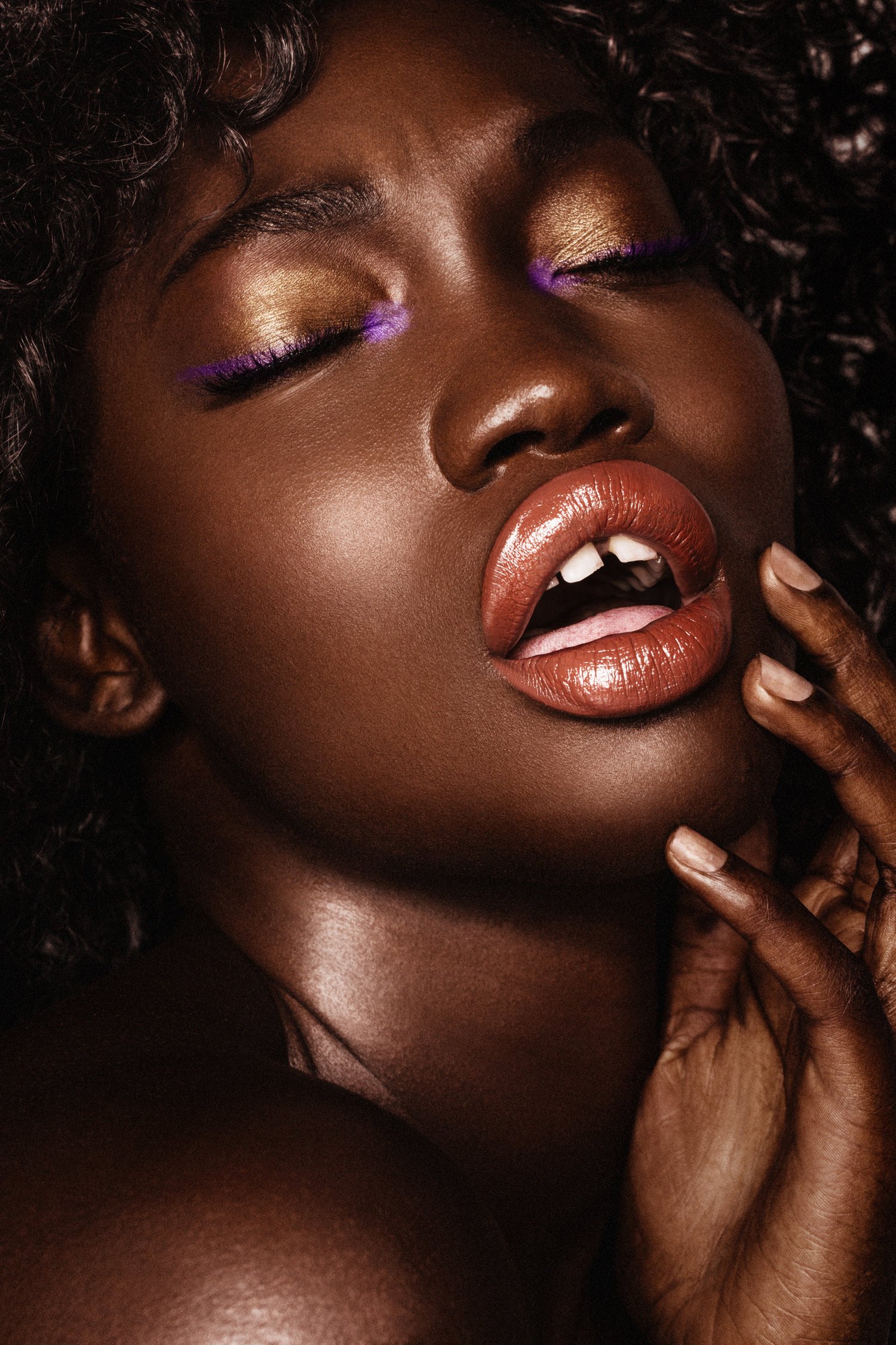 A close-up portrait of a woman with dark skin and curly hair, wearing gold eyeshadow, purple eyeliner, and glossy lipstick, touching her face with her fingers. Beauty Photographer Los Angeles.
