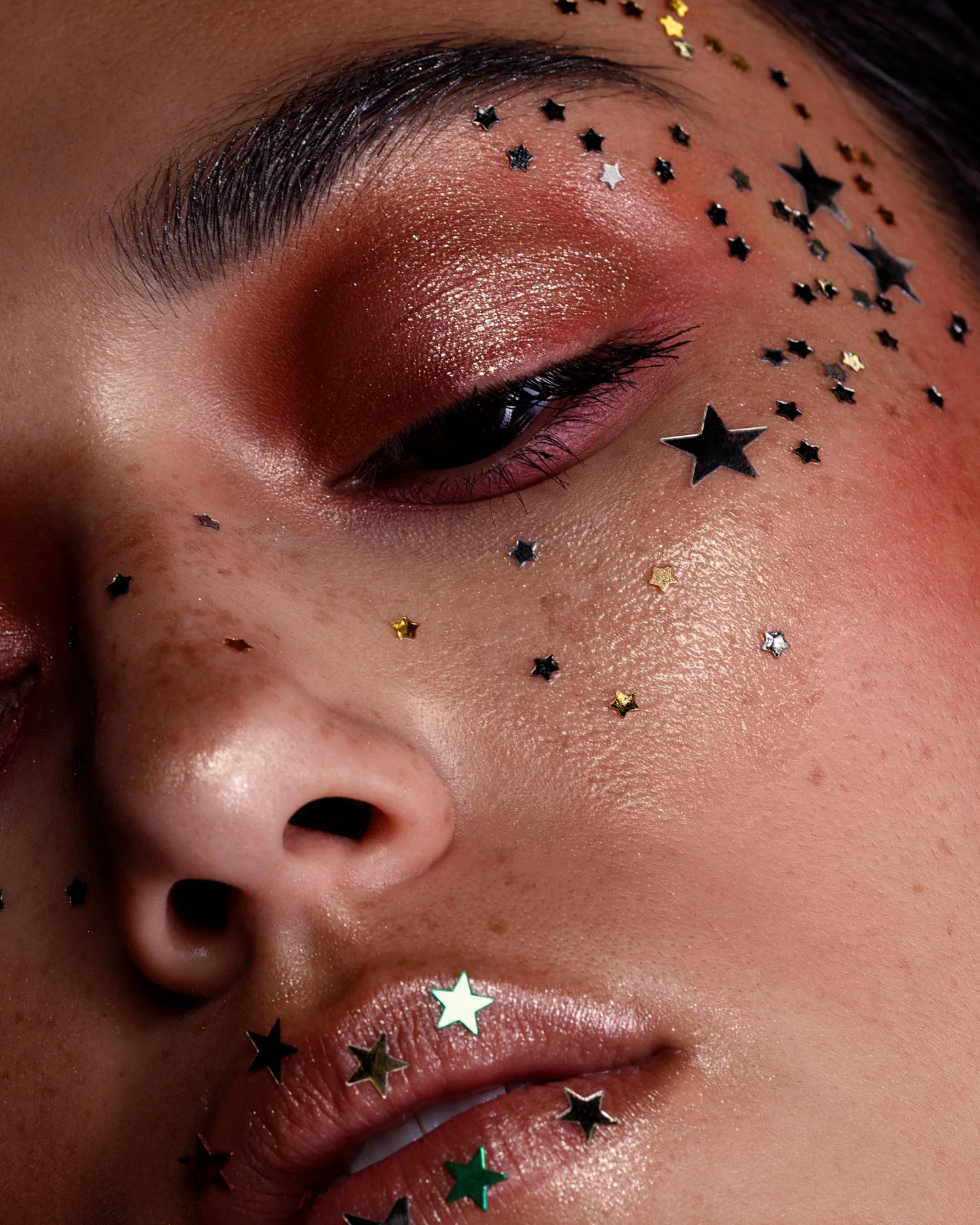 Close-up of a person's face with glittery makeup, star-shaped confetti on the face, and closed eye.