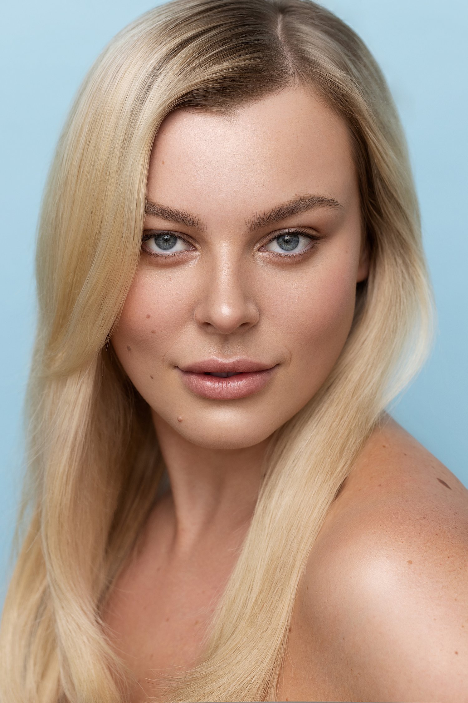 Close-up portrait of a young woman with long blonde hair, blue eyes, and light skin, smiling softly against a light blue background. Beauty Photographer Los Angeles.